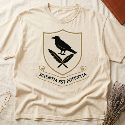 Ivory tee with cream-and-gold heraldic shield, black raven over crossed quills, scroll banner "SCIENTIA EST POTENTIA" below.