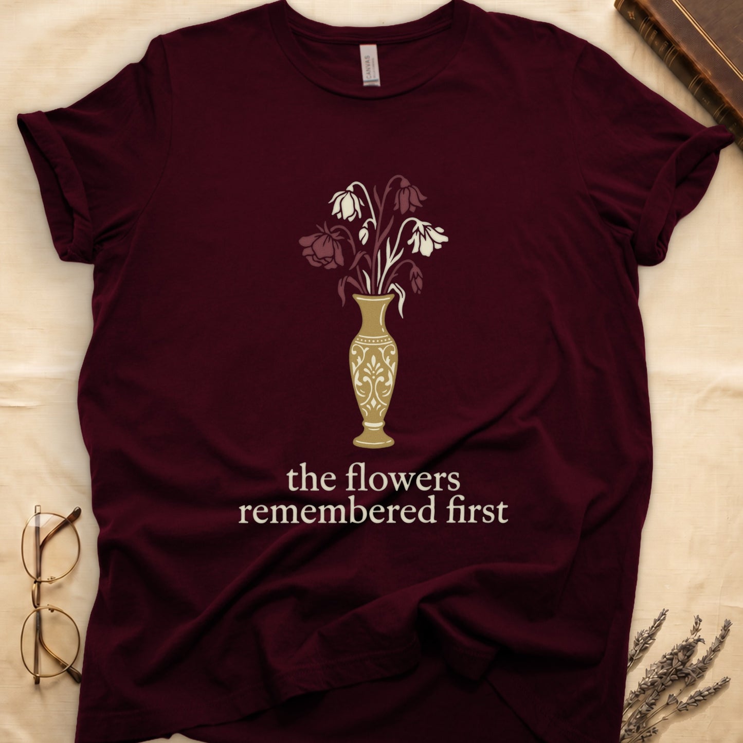 Maroon tee with gold ornate vase, wilting cream lilies and dark red roses, "the flowers remembered first" in cream serif.