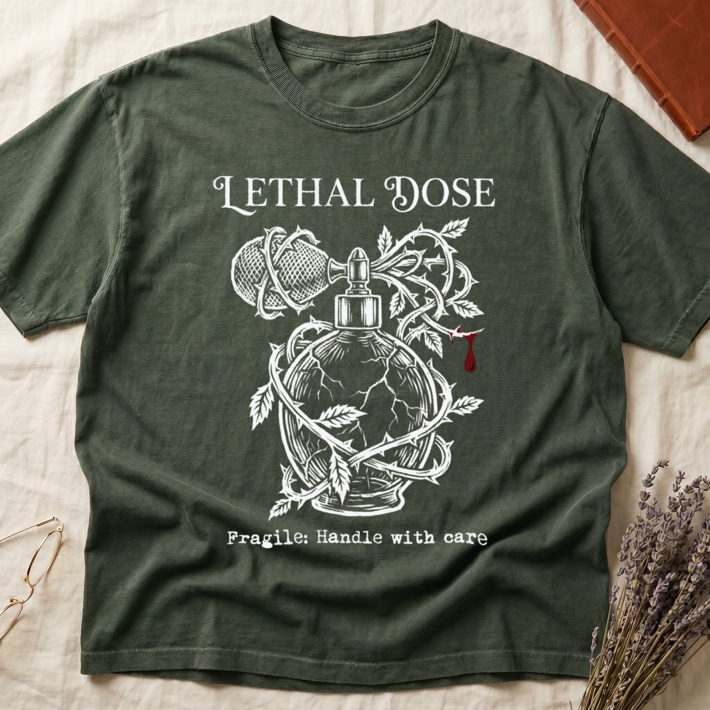 Green tee with cracked perfume bottle wrapped in thorns, blood drop, "LETHAL DOSE" top, "Fragile: Handle with care" below.