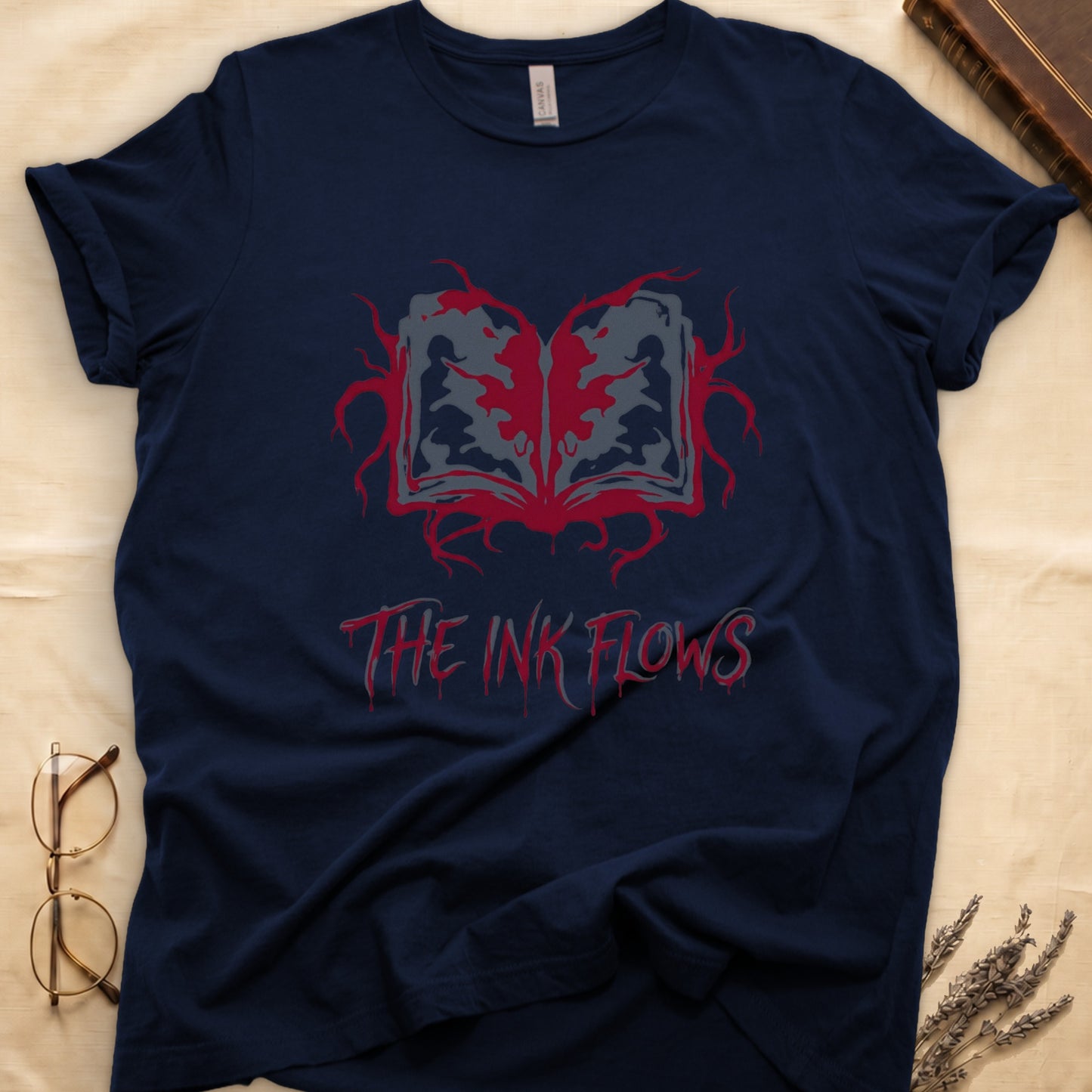 Navy tee with open book bleeding red ink into a gray face on green ground, "THE INK FLOWS" in dripping serif.