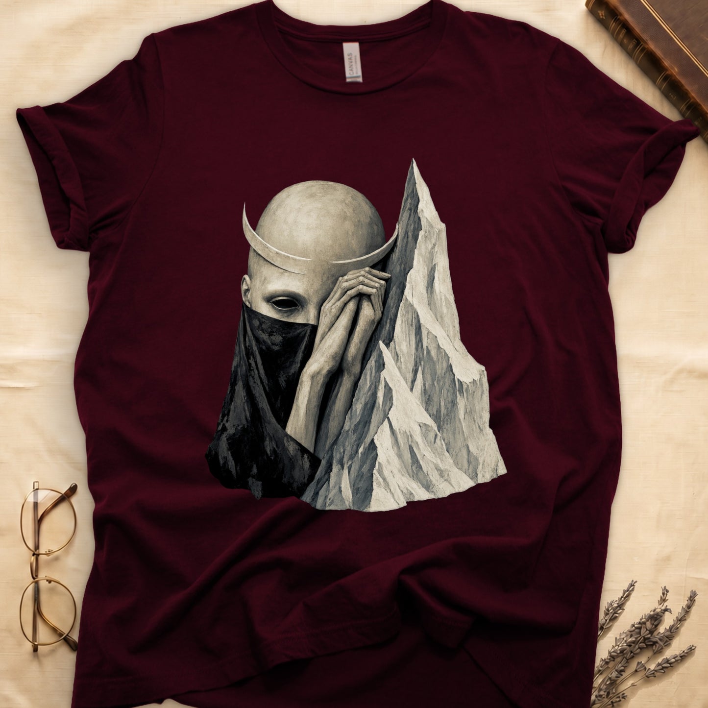 Maroon t-shirt with a graphic of a bald, pale figure wearing a dark veil and crescent horns next to a mountain.