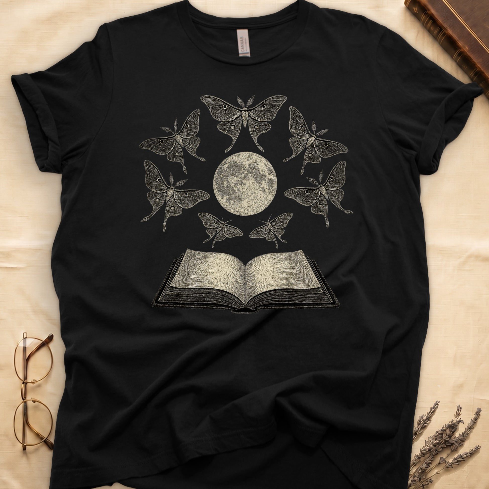 Black tee with full moon center, seven luna moths in halo around, open book below, cream-on-black engraving.