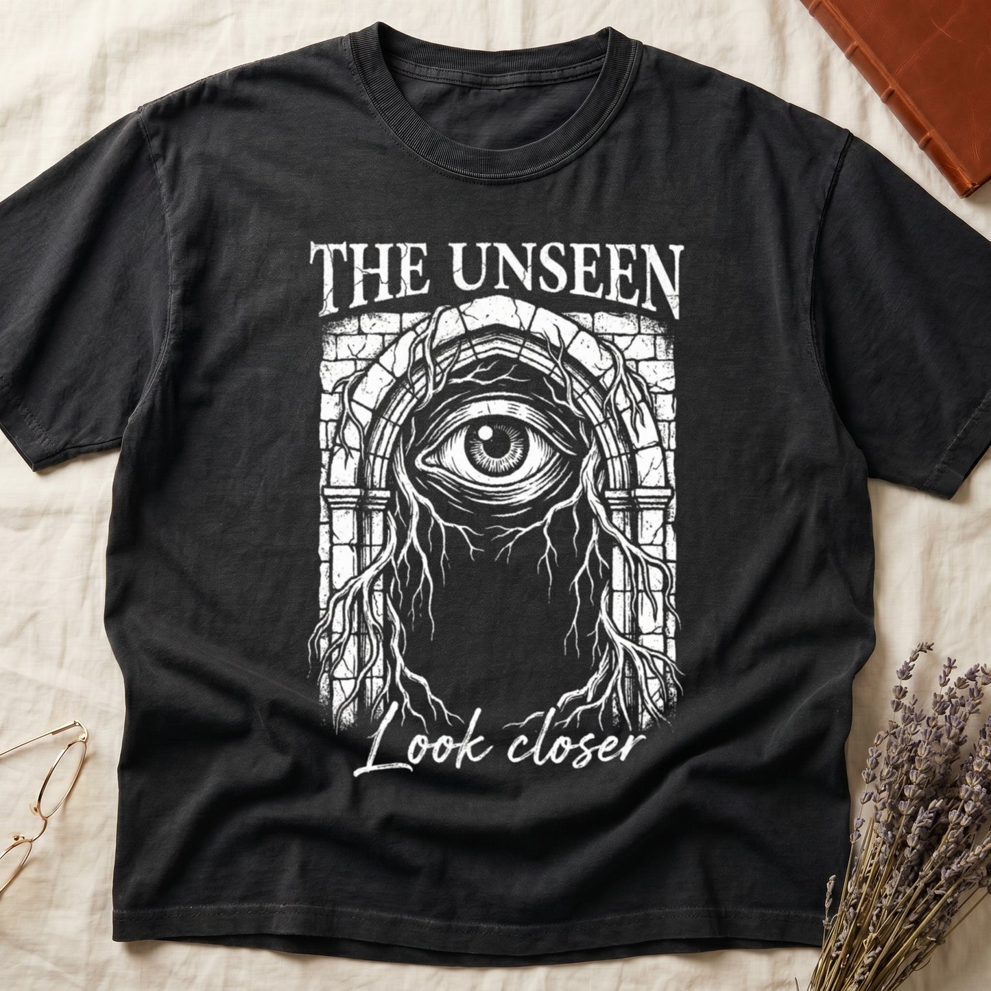 Black tee with Gothic stone arch, open eye, roots trailing, "THE UNSEEN" top, "Look closer" cursive below.