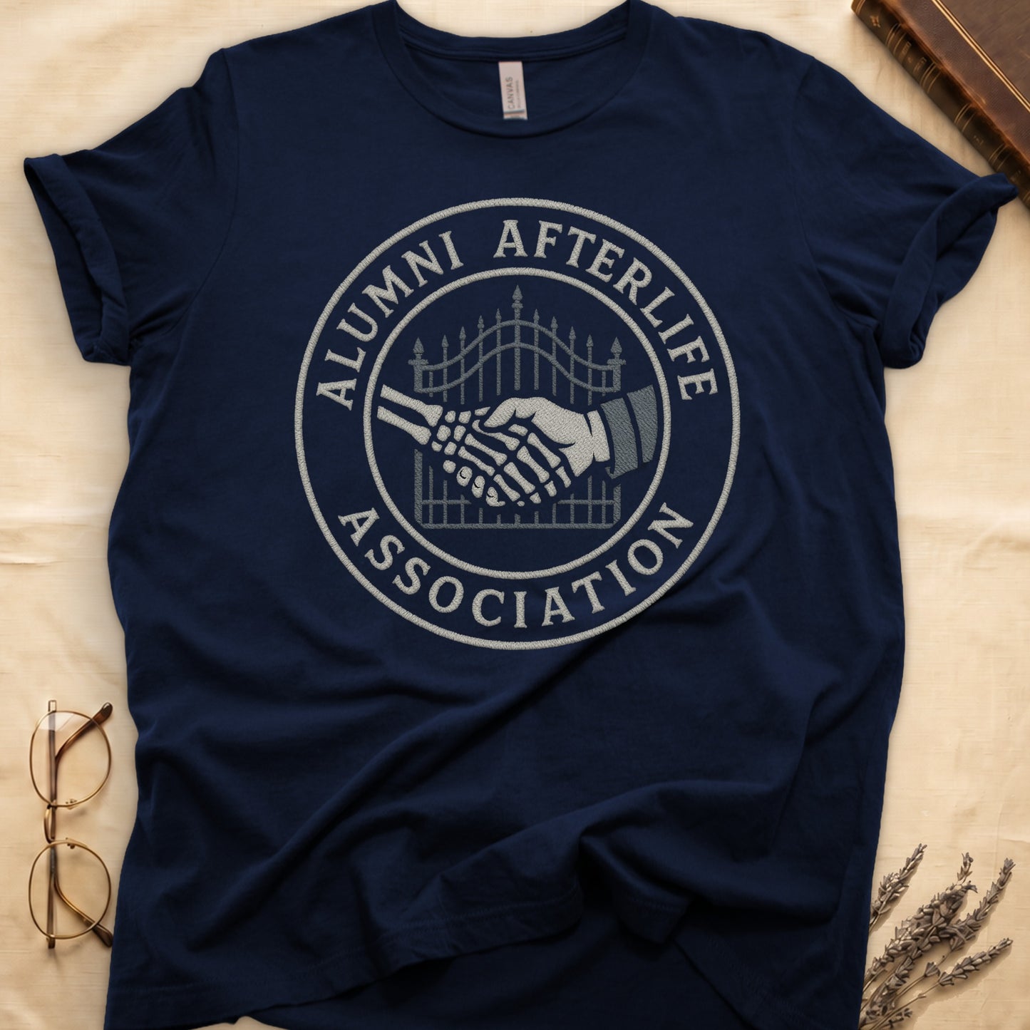 Black tee with embroidered patch, skeletal handshake at cemetery gate, "ALUMNI AFTERLIFE" top, "ASSOCIATION" bottom.