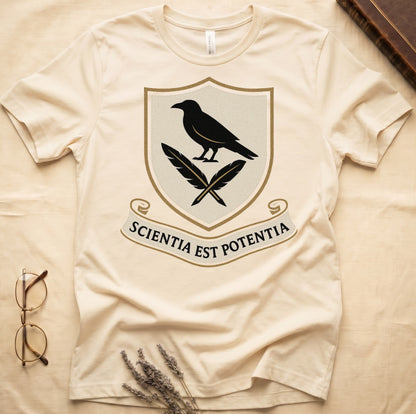 Natural tee with cream-and-gold heraldic shield, black raven over crossed quills, scroll banner "SCIENTIA EST POTENTIA" below.
