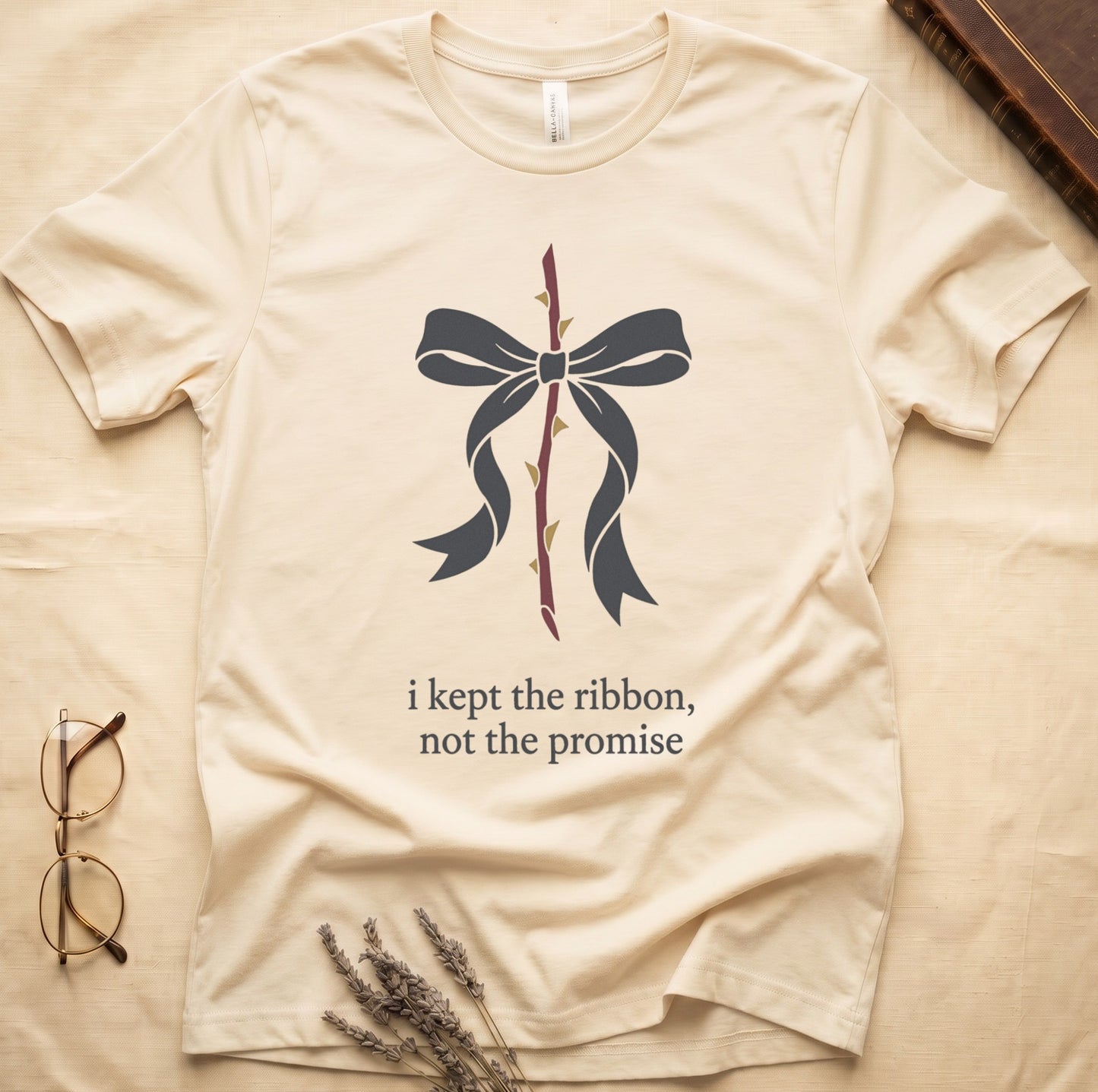 Natural tee with cream bow tied around thorny rose stem, "i kept the ribbon, not the promise" in cream serif below.