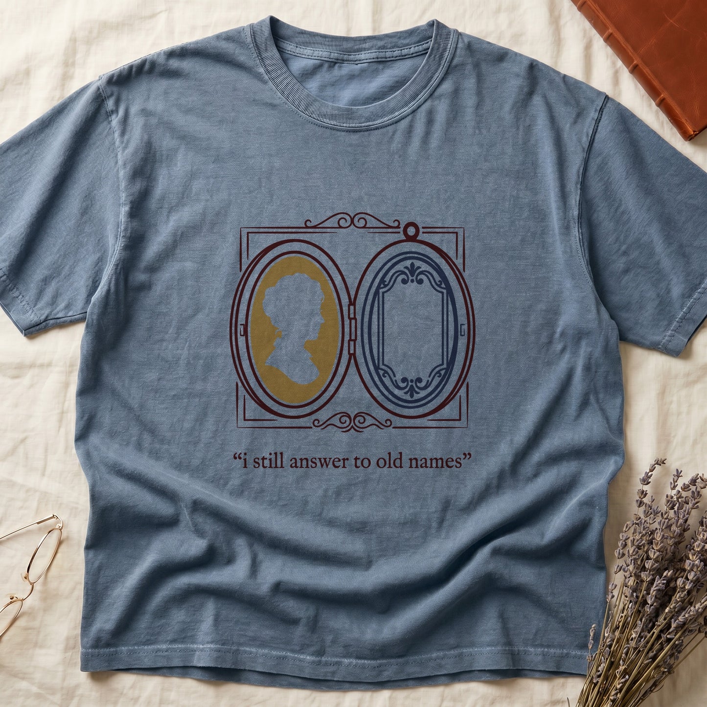 Blue Jean tee with Victorian double-locket, silhouette portrait in gold frame, "i still answer to old names" cream serif below.