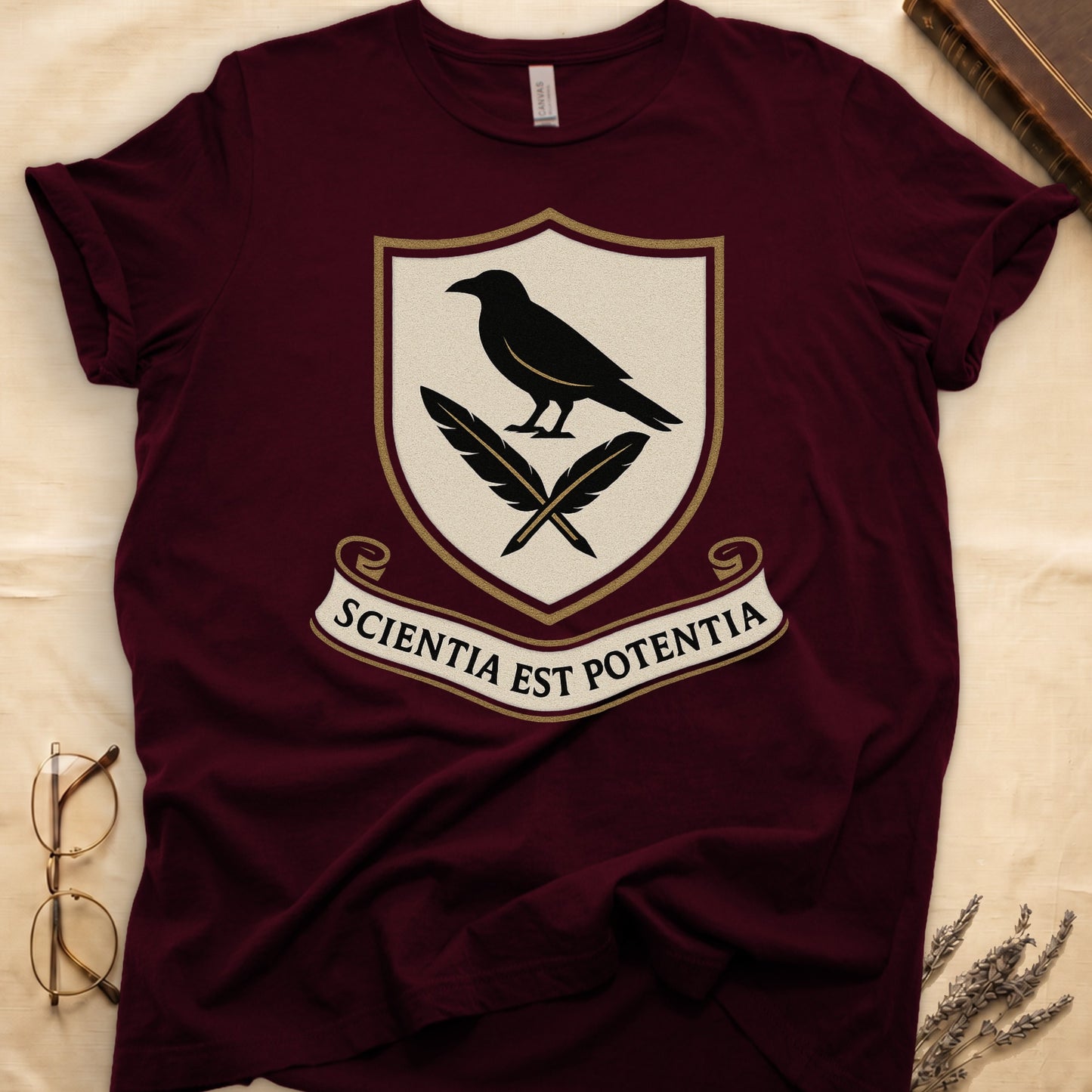 Maroon tee with cream-and-gold heraldic shield, black raven over crossed quills, scroll banner "SCIENTIA EST POTENTIA" below.