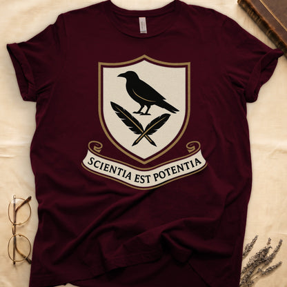 Maroon tee with cream-and-gold heraldic shield, black raven over crossed quills, scroll banner "SCIENTIA EST POTENTIA" below.