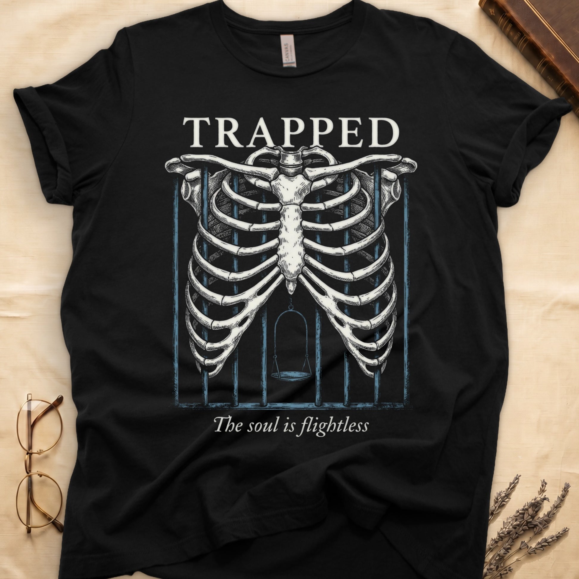 Black tee with anatomical ribcage on prison bars, bird swing inside, "TRAPPED" top, "The soul is flightless" italic below.
