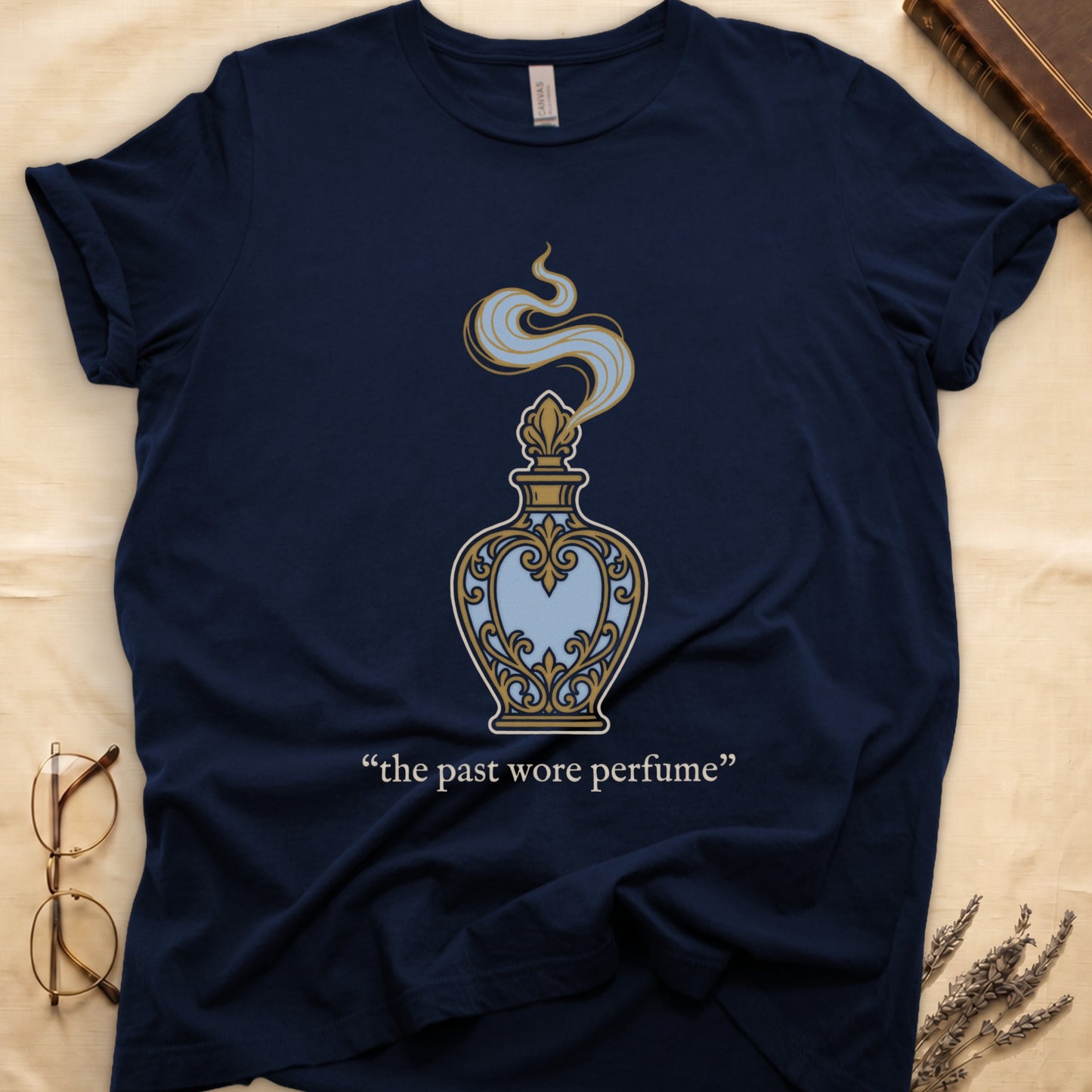 Navy tee with ornate blue and gold perfume bottle, rising scent swirl, "the past wore perfume" cream serif below.