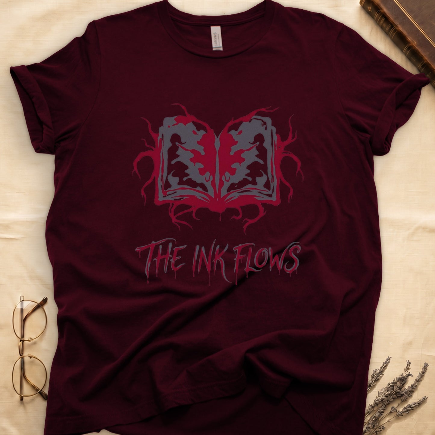 Maroon tee with open book bleeding red ink into a gray face on green ground, "THE INK FLOWS" in dripping serif.