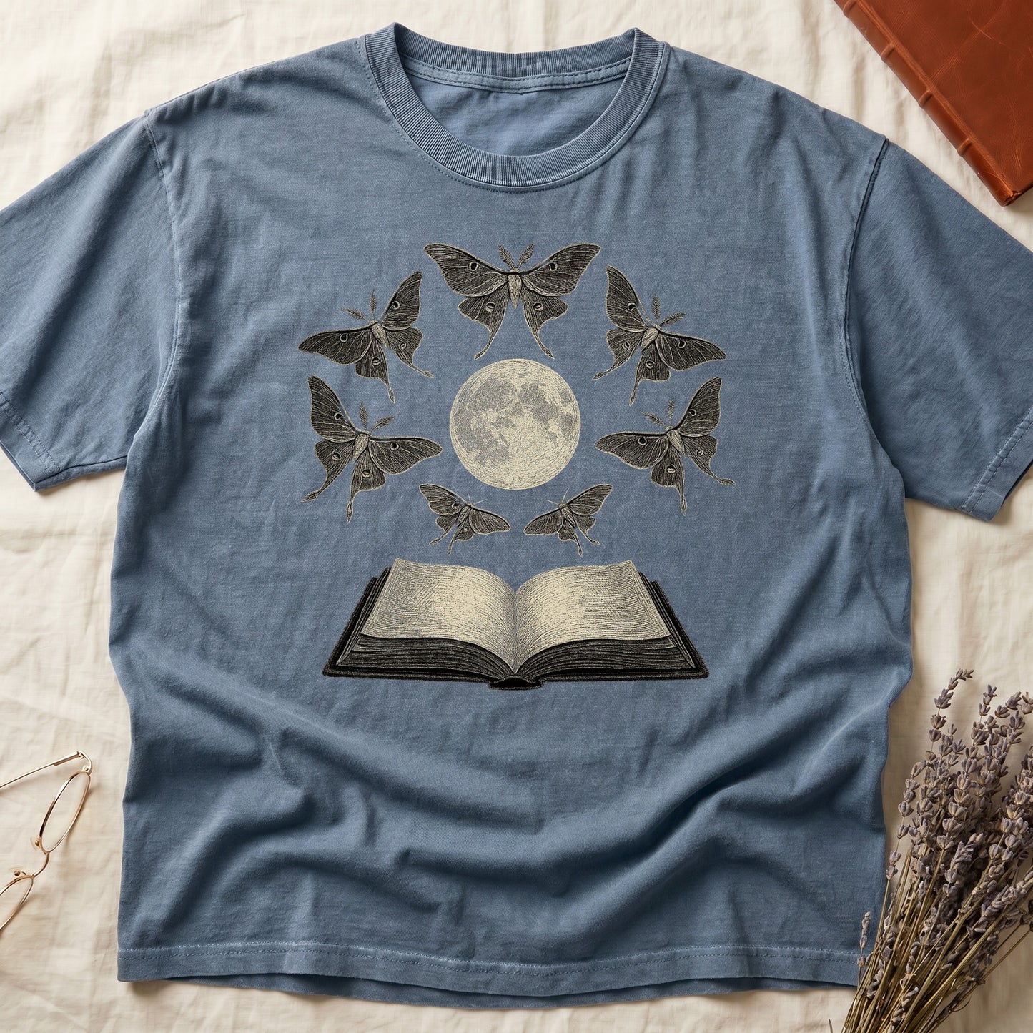 Blue Jean tee with full moon center, seven luna moths in halo around, open book below, cream-on-black engraving.