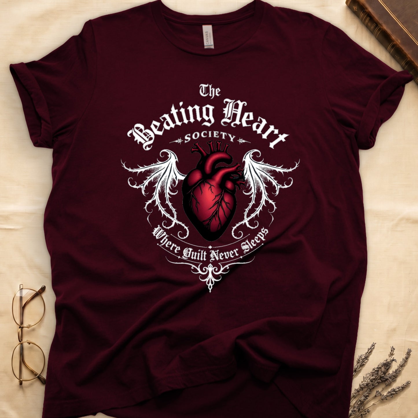 Maroon tee with anatomical red heart, gothic wings, "The Beating Heart SOCIETY," "Where Guilt Never Sleeps" below.