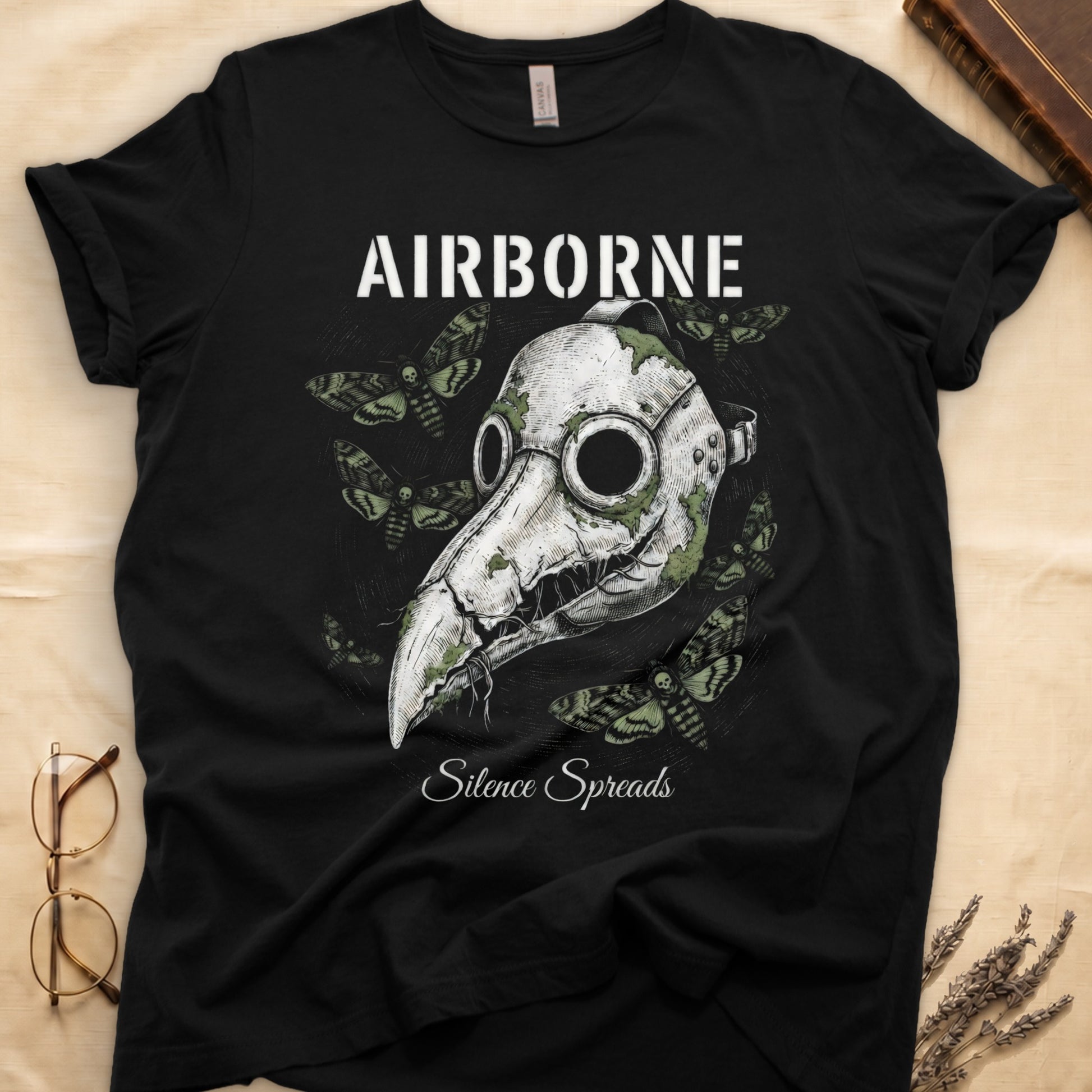 Black tee with plague doctor mask in cream linework, death's-head moths, "AIRBORNE" top, "Silence Spreads" cursive below.