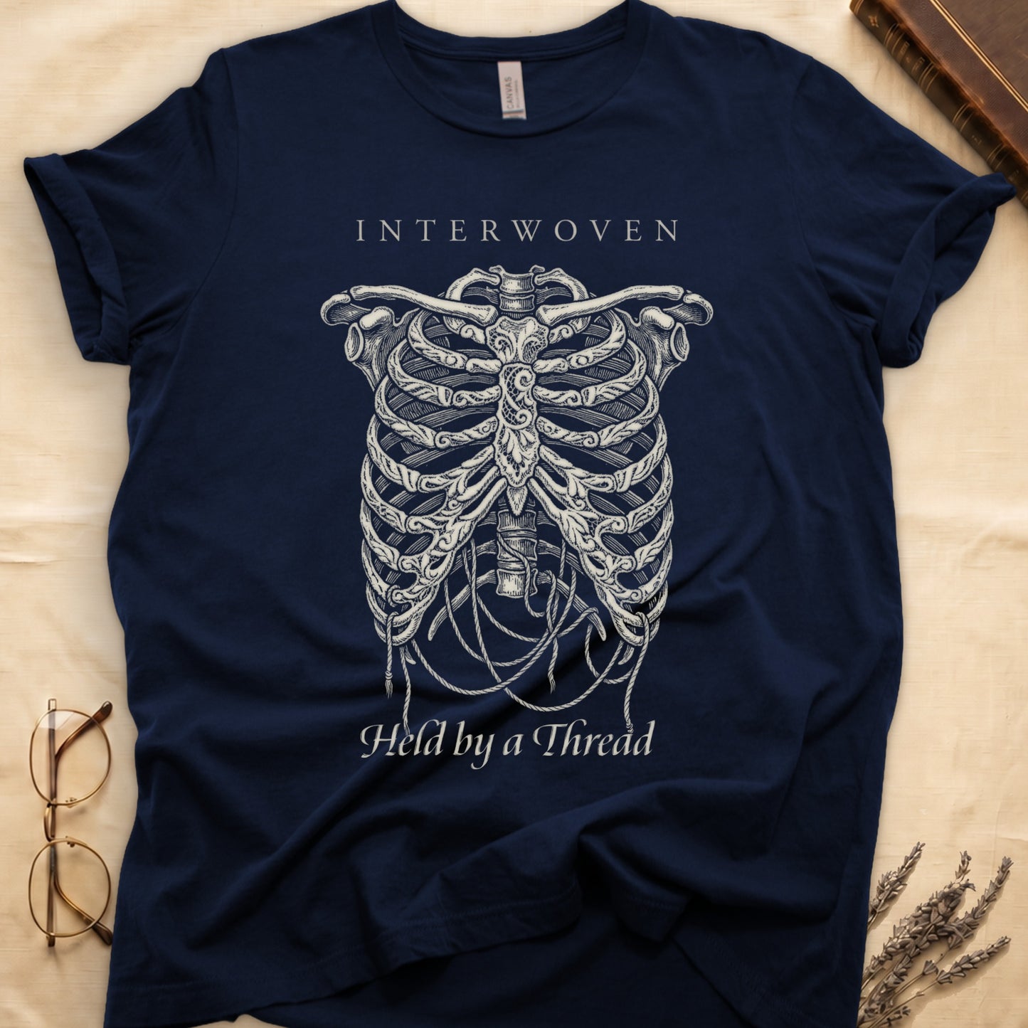 Navy tee with cream skeletal ribcage suspended by trailing threads, "Held by a Thread" in serif on plate below.