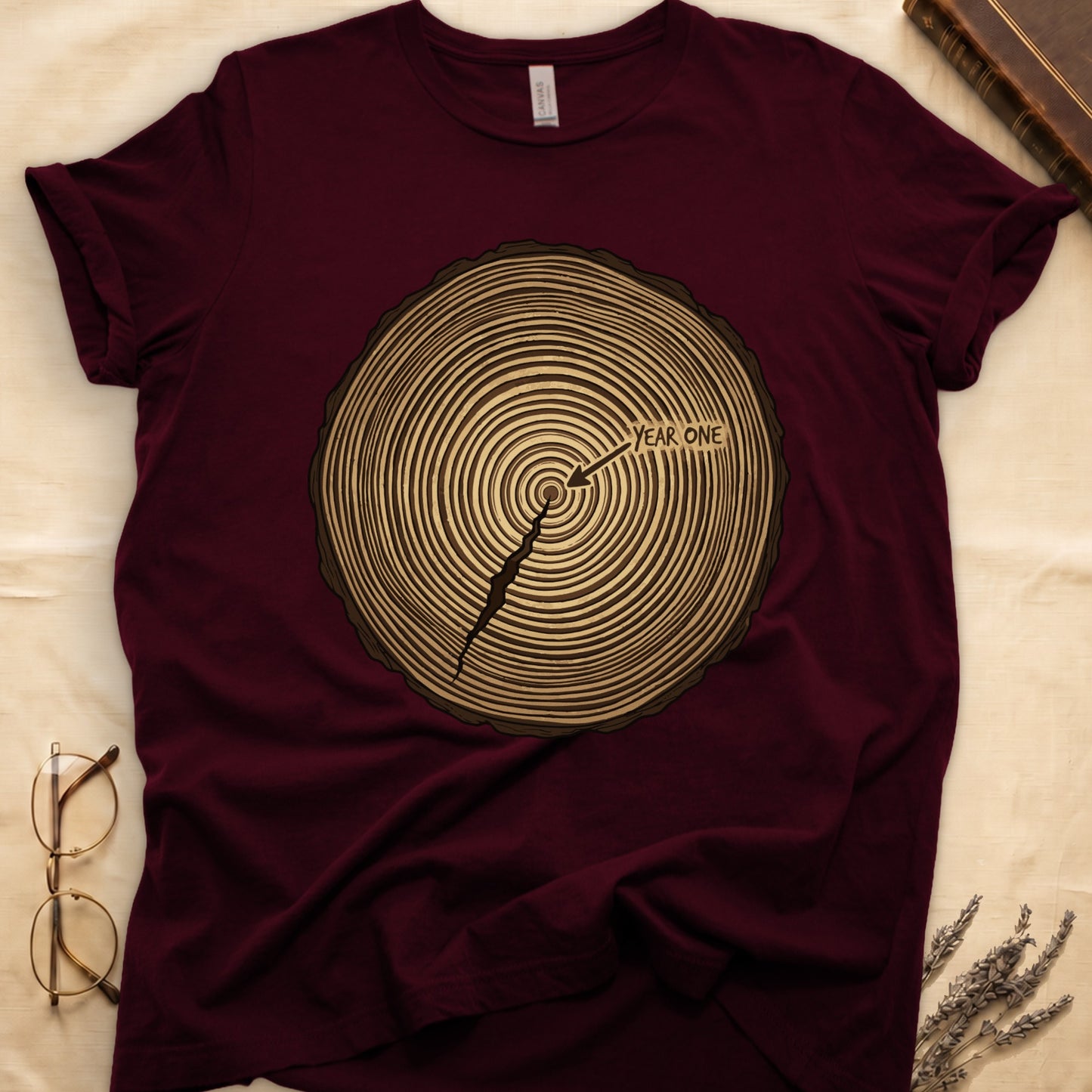 Maroon tee with cross-section of tree rings in warm brown, arrow pointing to center labeled "YEAR ONE" in serif type.