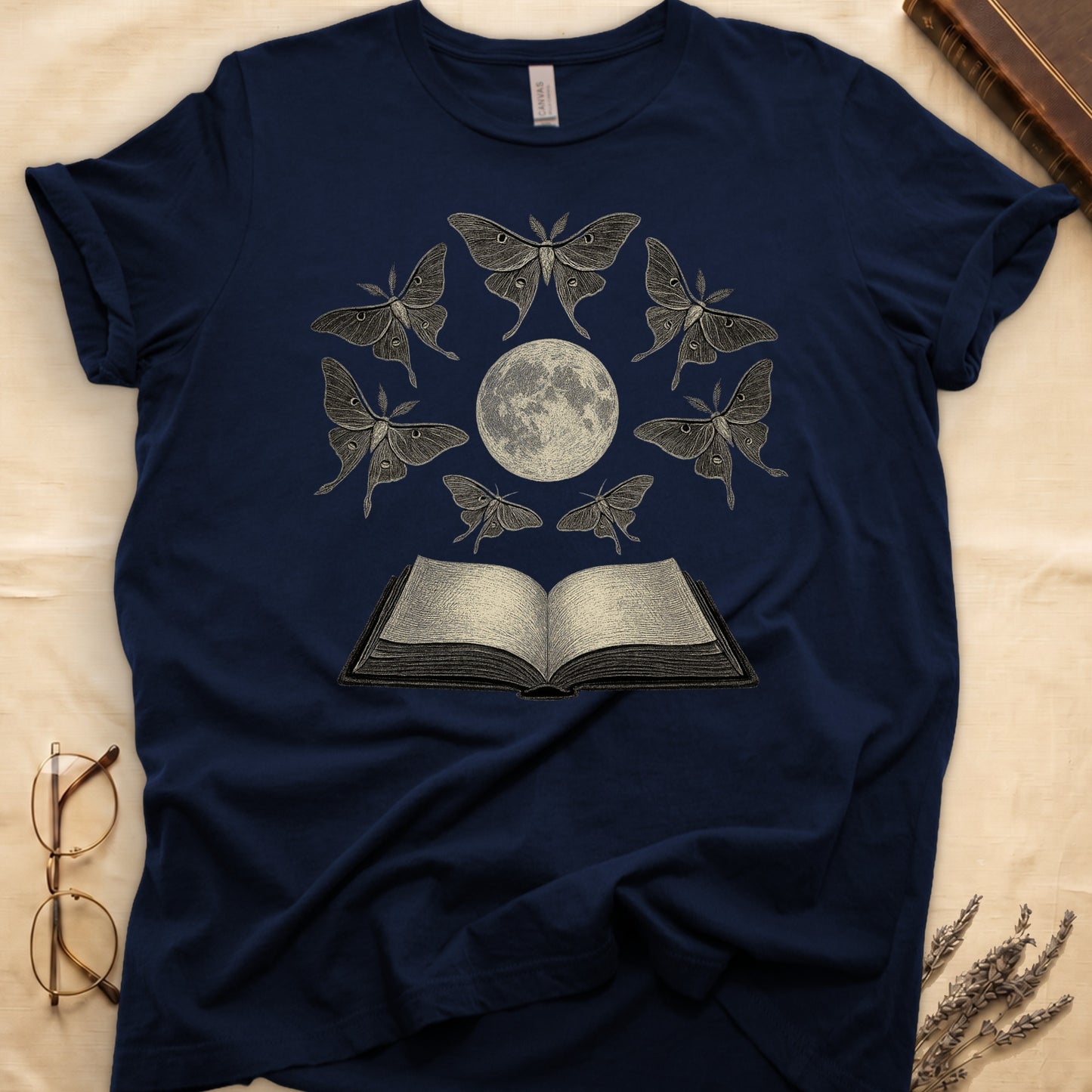 Navy tee with full moon center, seven luna moths in halo around, open book below, cream-on-black engraving.