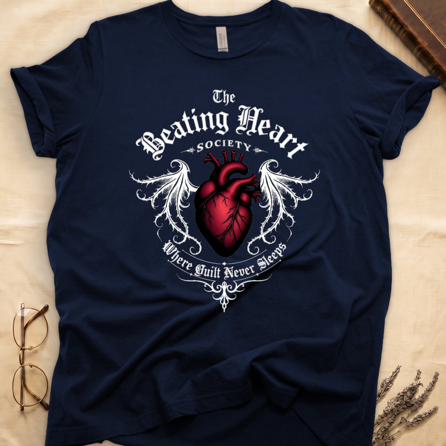 Navy tee with anatomical red heart, gothic wings, "The Beating Heart SOCIETY," "Where Guilt Never Sleeps" below.