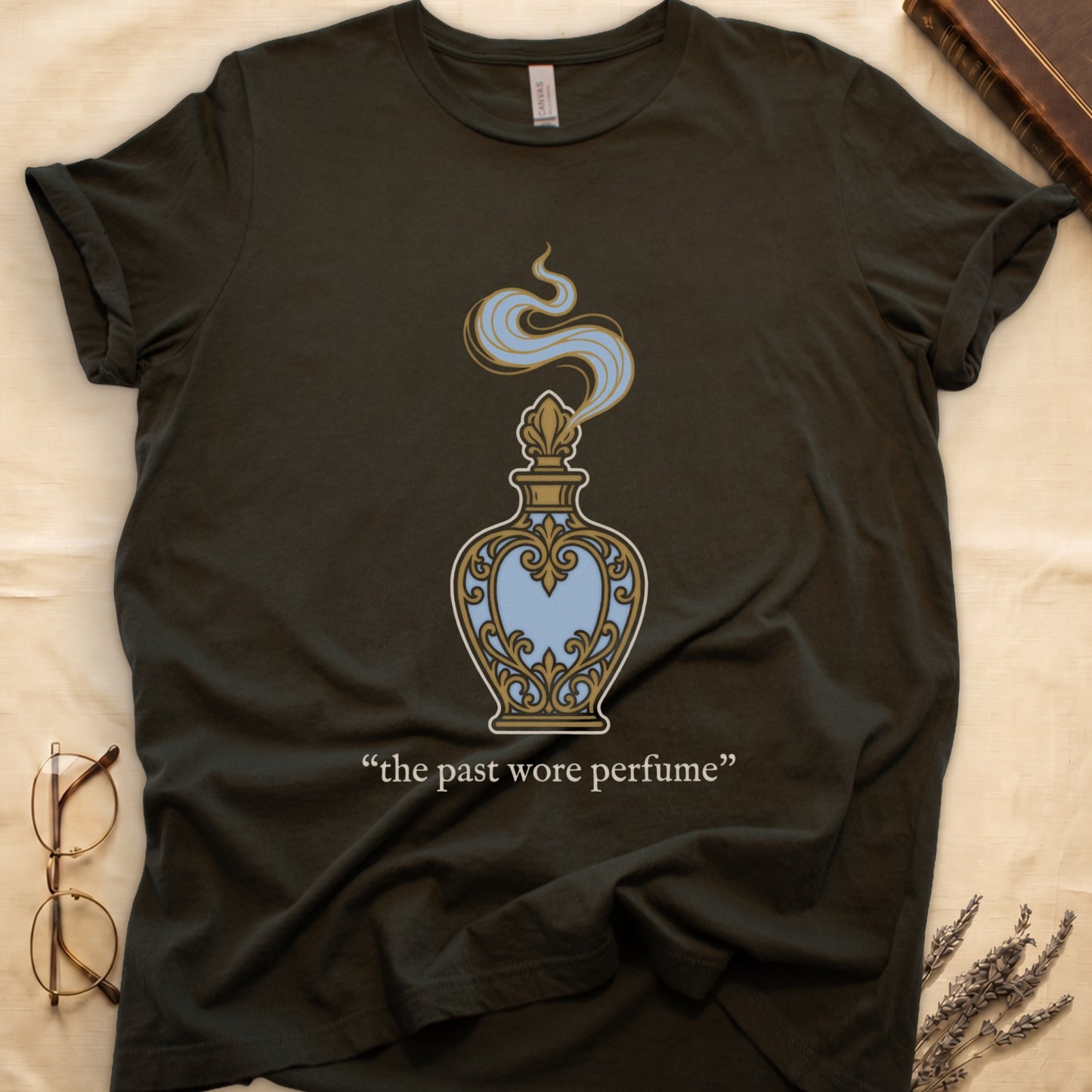 Military Green tee with ornate blue and gold perfume bottle, rising scent swirl, "the past wore perfume" cream serif below.