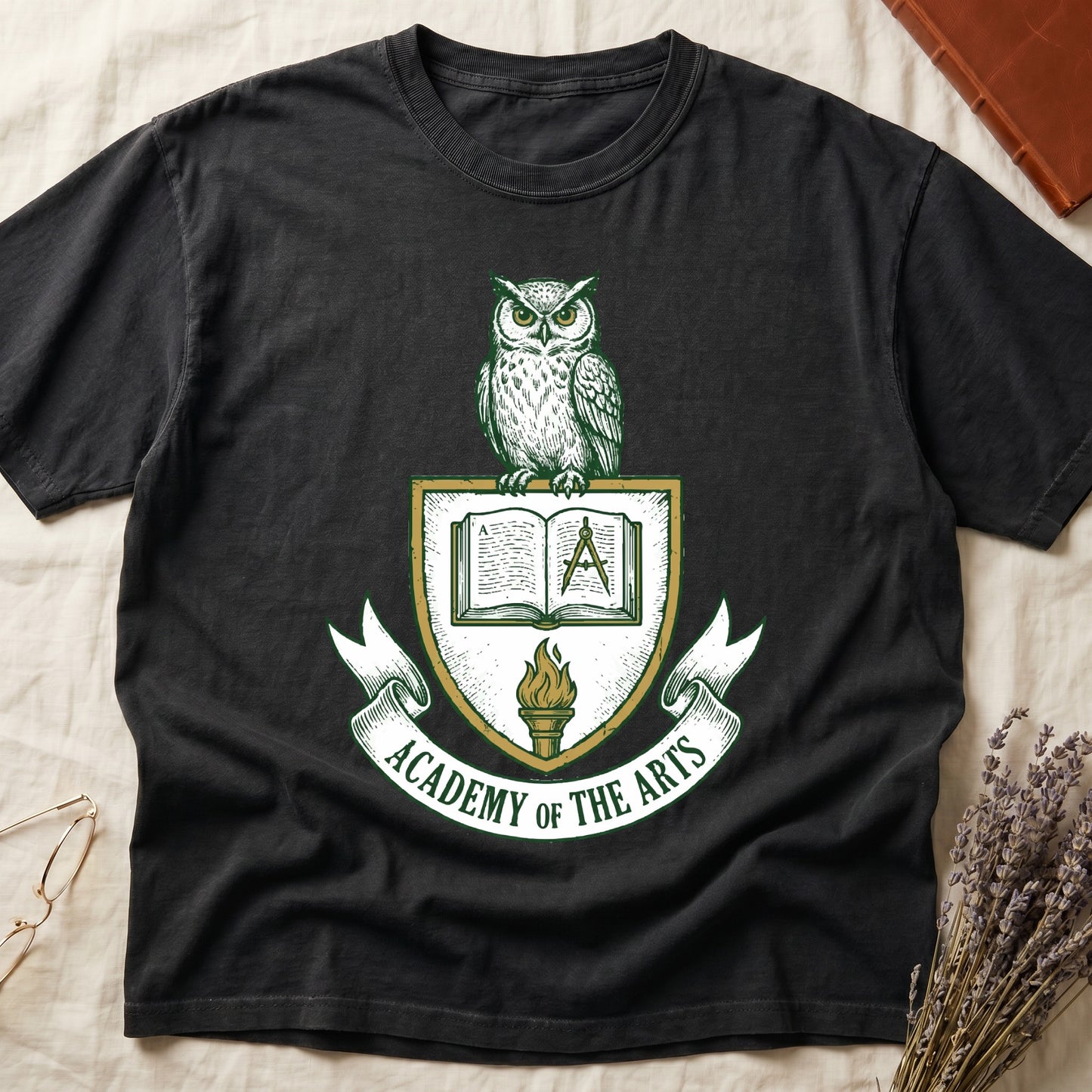 Black tee with heraldic shield crest, owl perched on top, open book and dividers center, "ACADEMY OF THE ARTS" on a banner.