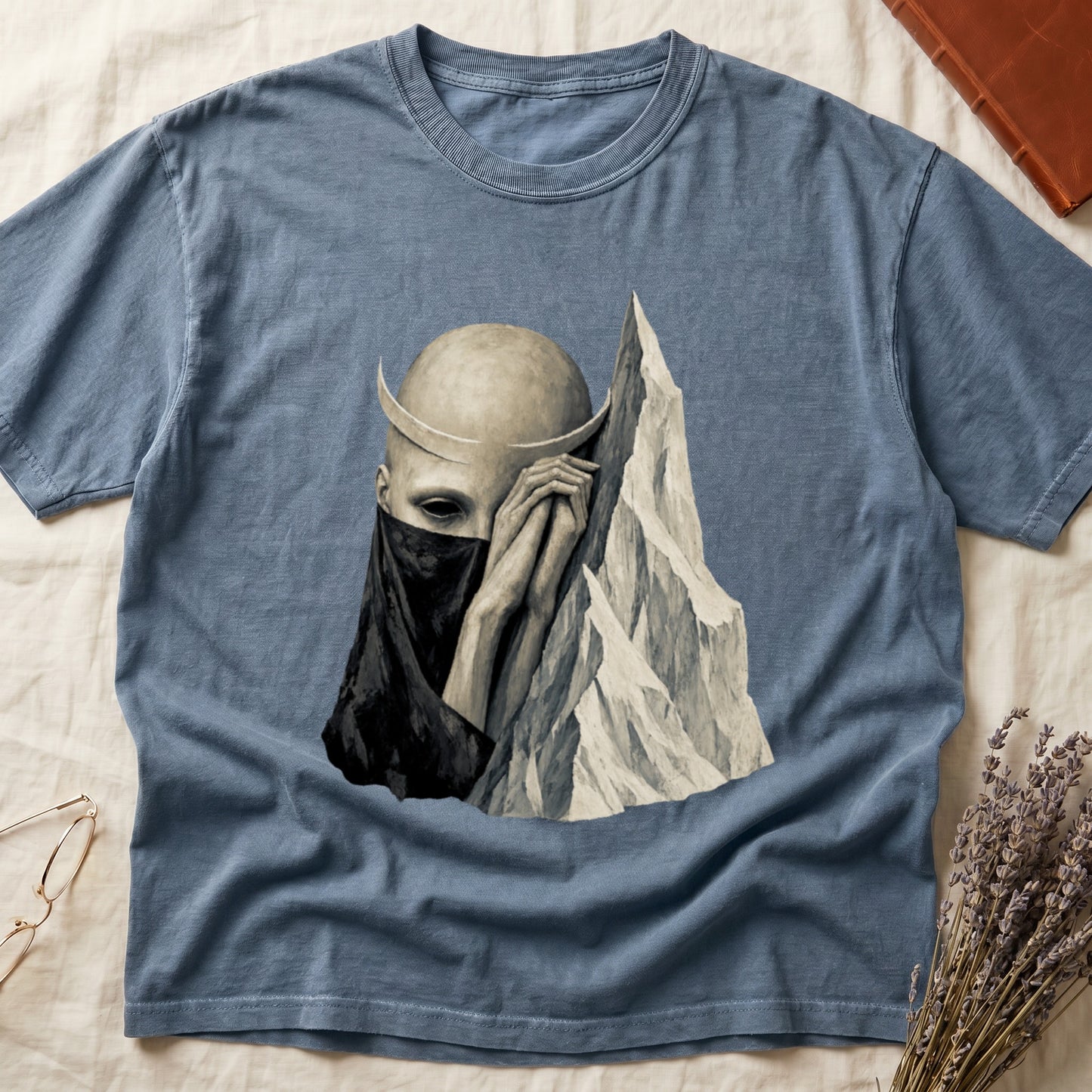 Blue t-shirt with a dark figure wearing a veil and crescent moon horns next to mountains.