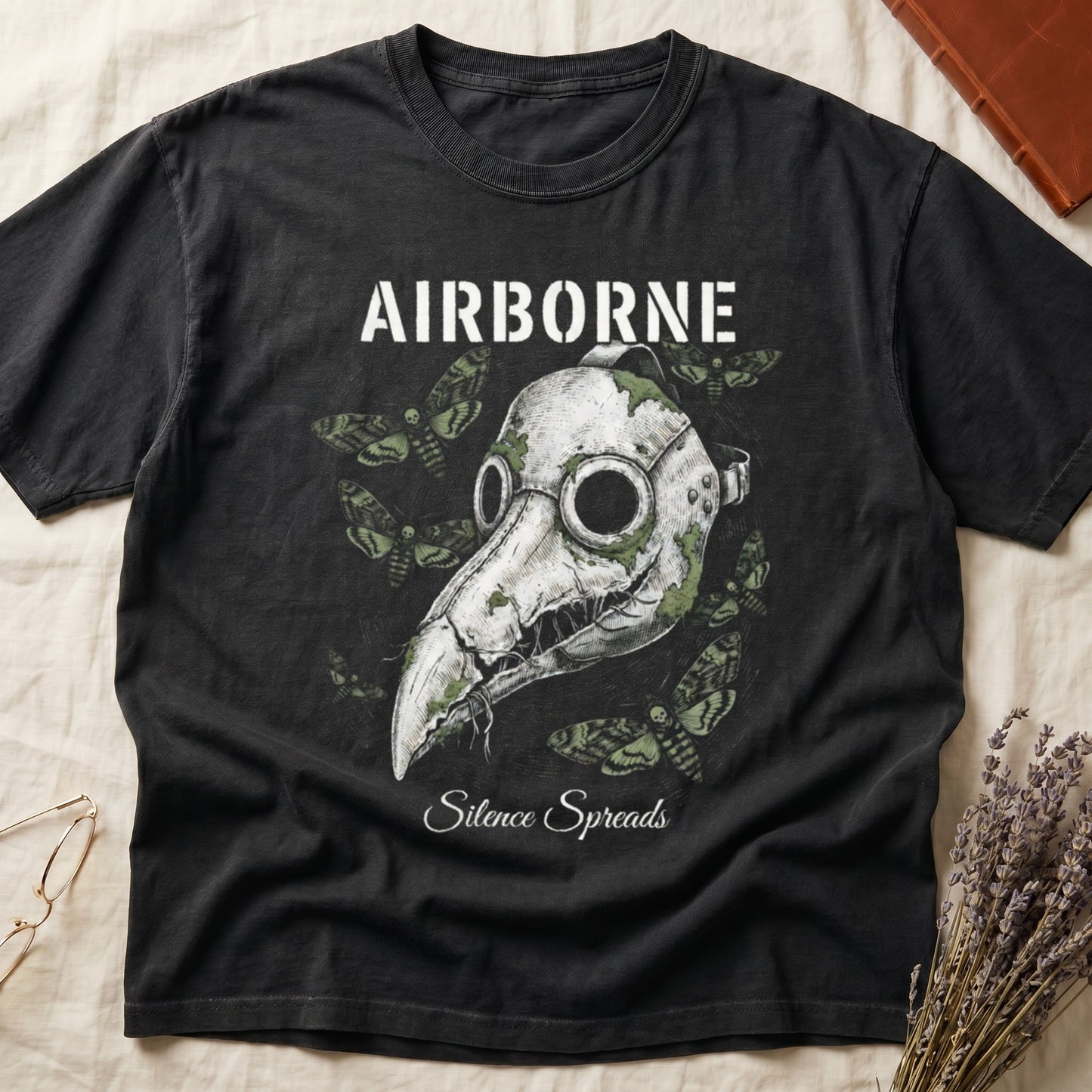 Black tee with plague doctor mask in cream linework, death's-head moths, "AIRBORNE" top, "Silence Spreads" cursive below.