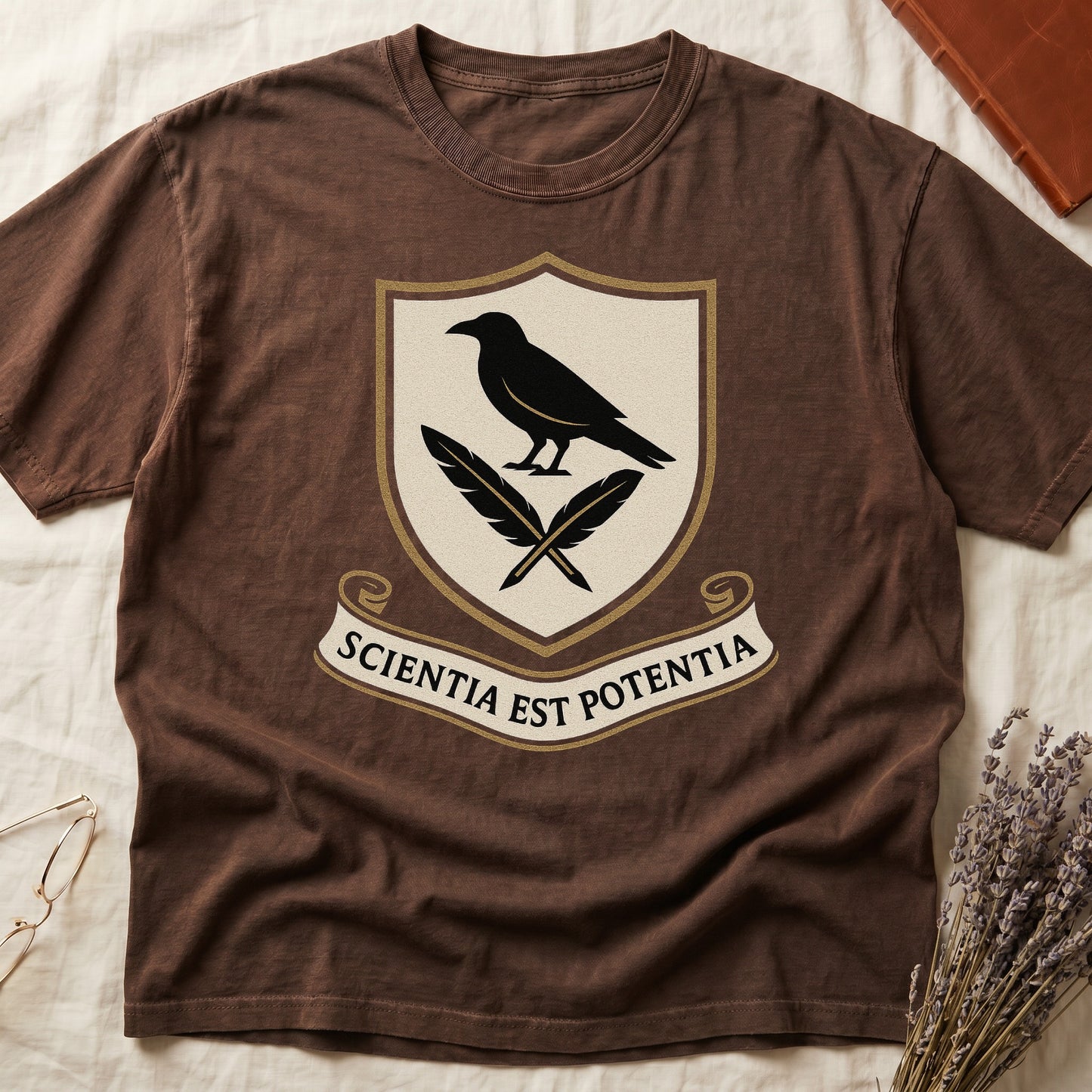 Espresso tee with cream-and-gold heraldic shield, black raven over crossed quills, scroll banner "SCIENTIA EST POTENTIA" below.