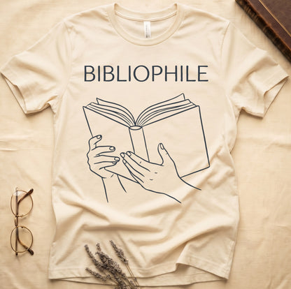 Natural tee with two hands cradling an open book in cream and brown linework, "BIBLIOPHILE" in serif capitals above.