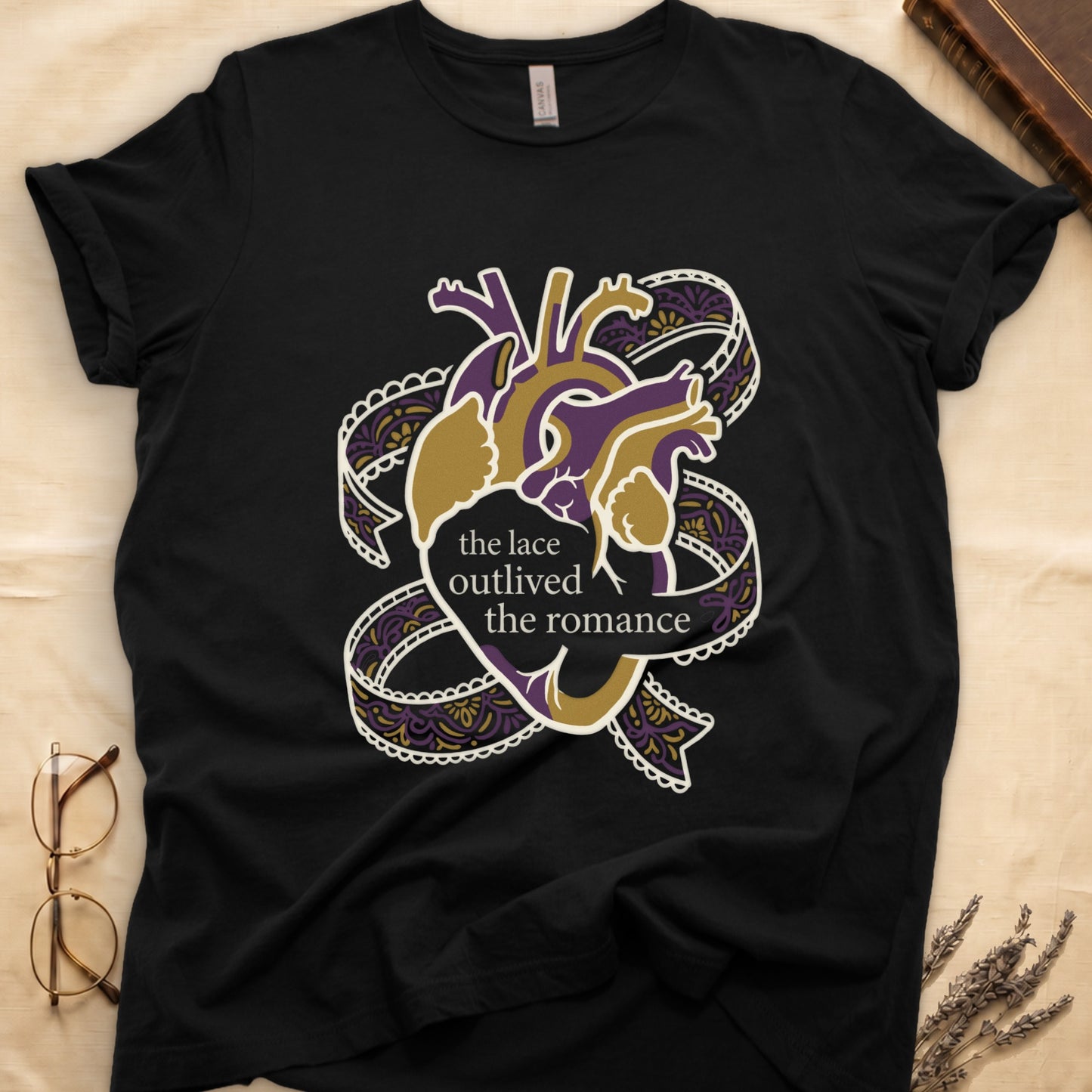 Black tee with anatomical heart in purple and gold wrapped in lace ribbons, "the lace outlived the romance" in serif.