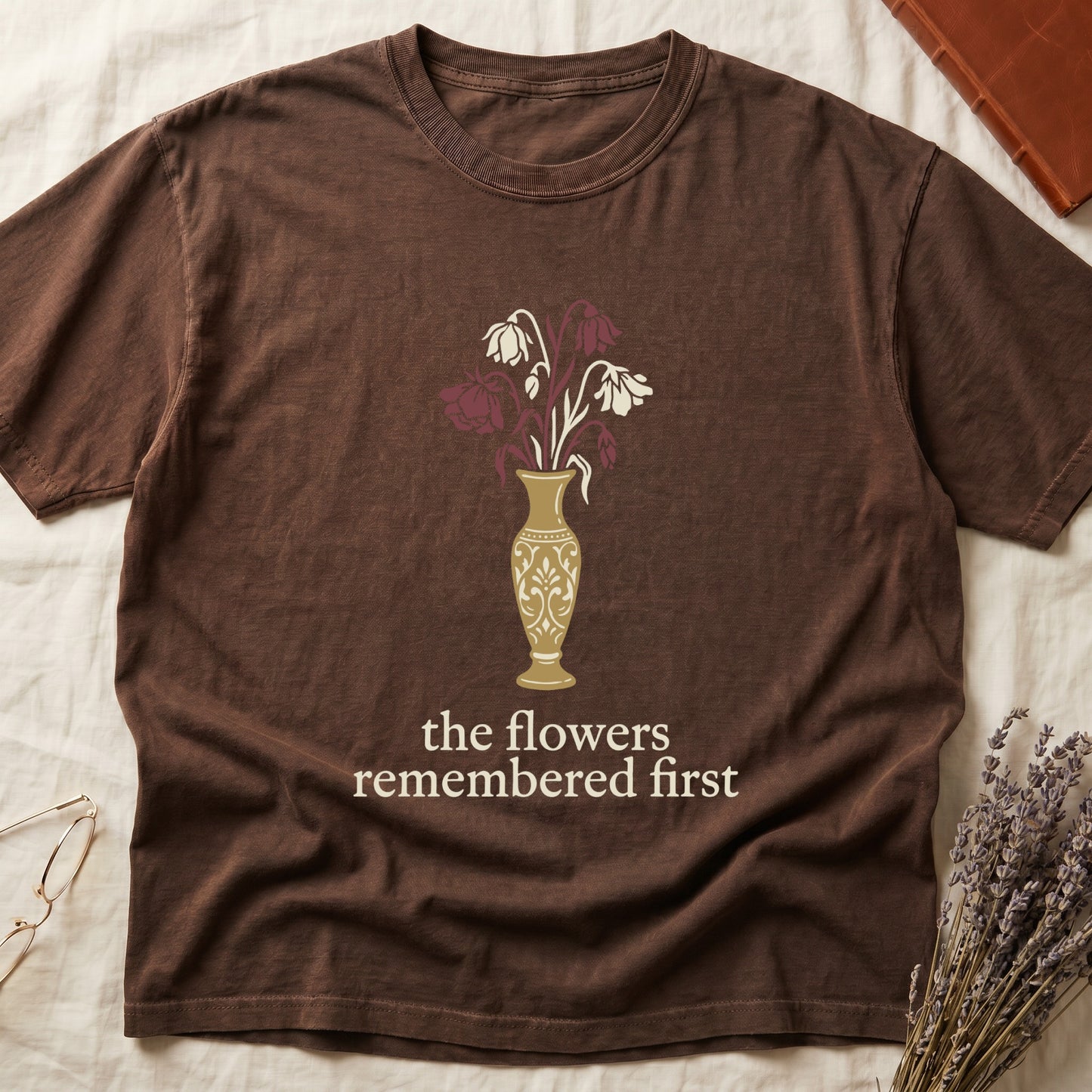 Espresso tee with gold ornate vase, wilting cream lilies and dark red roses, "the flowers remembered first" in cream serif.