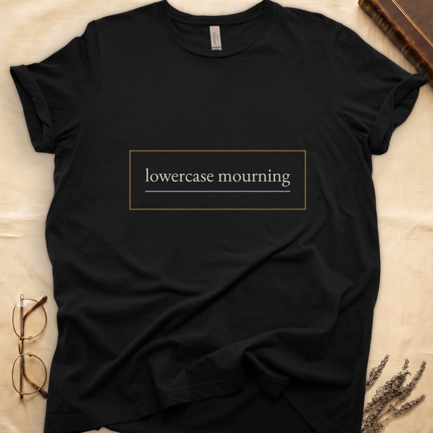 Black tee with "lowercase mourning" in cream serif inside gold rectangle frame, lavender line below.