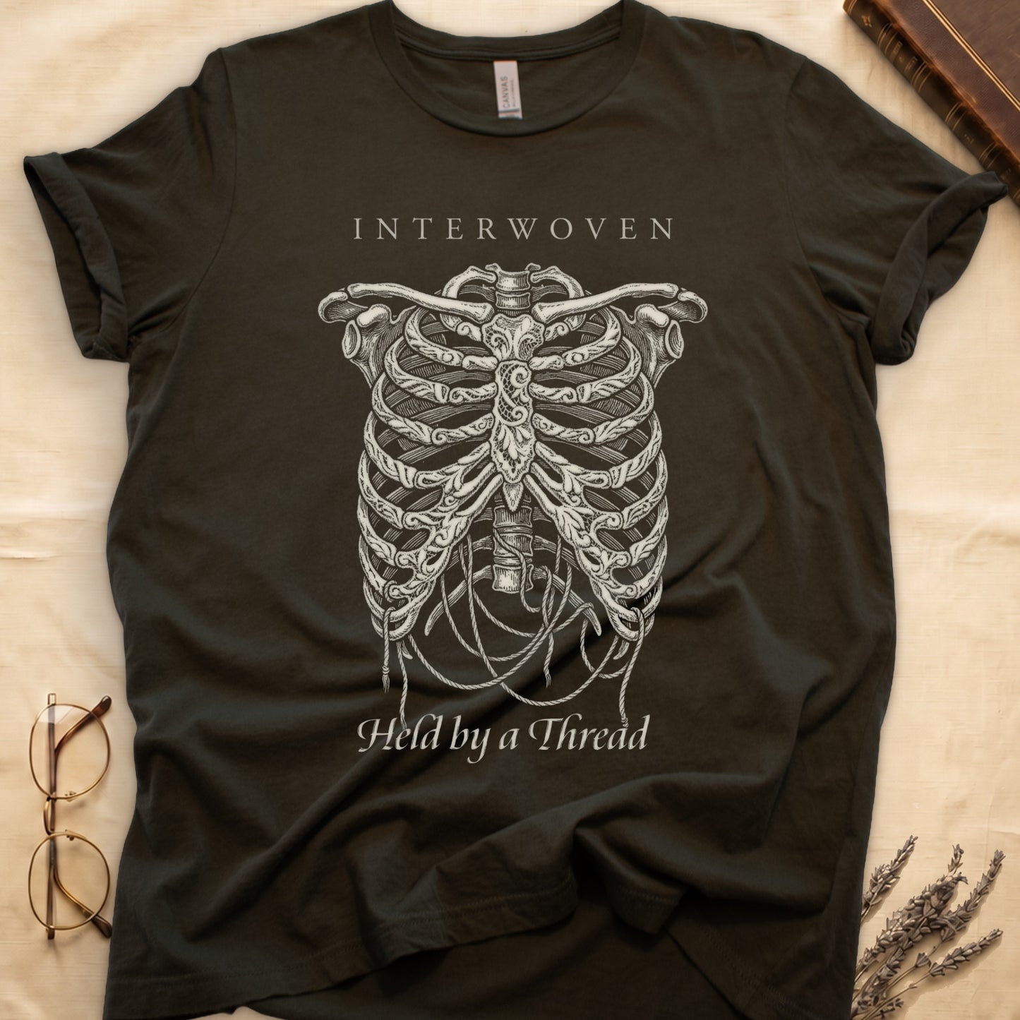 Military Green tee with cream skeletal ribcage suspended by trailing threads, "Held by a Thread" in serif on plate below.