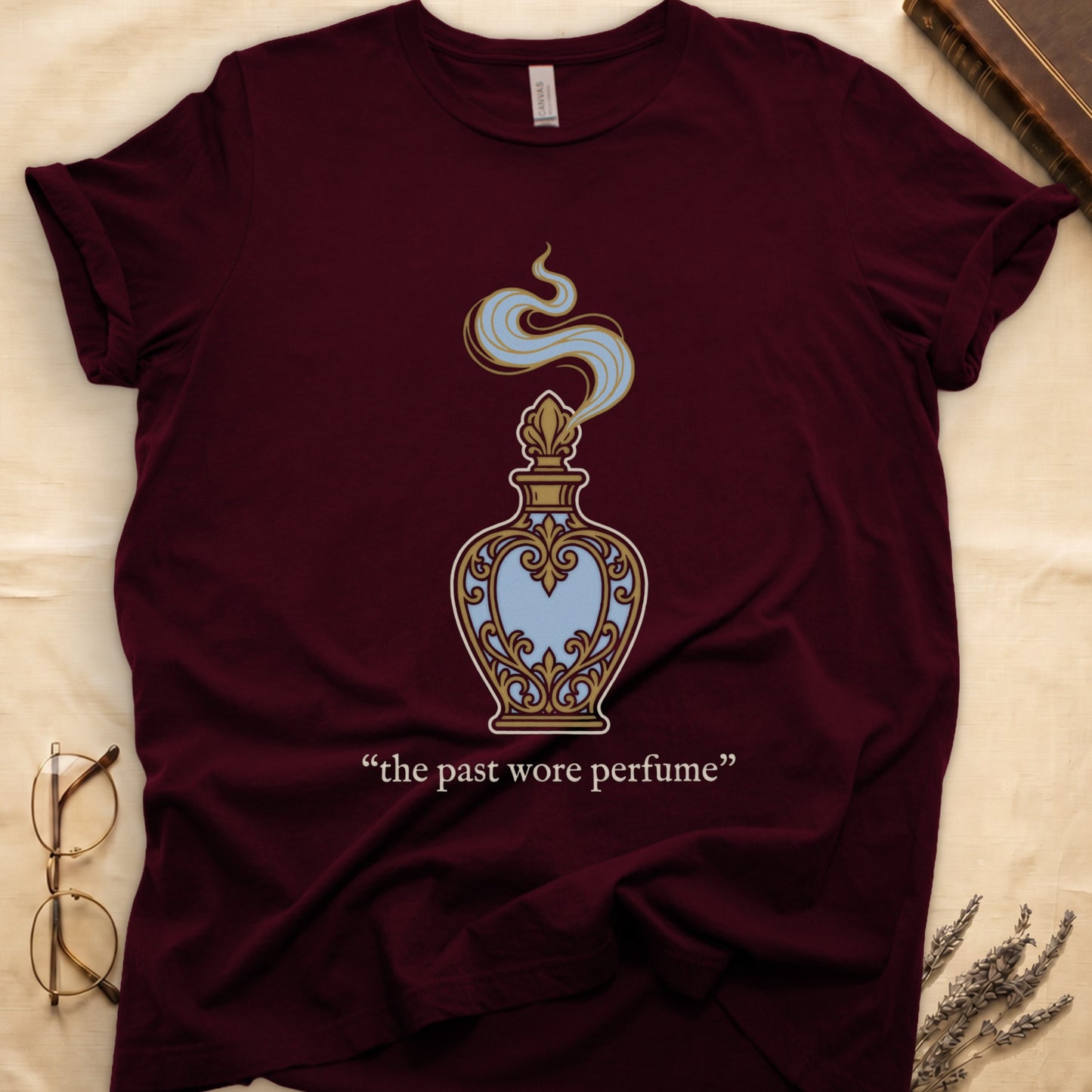 Maroon tee with ornate blue and gold perfume bottle, rising scent swirl, "the past wore perfume" cream serif below.