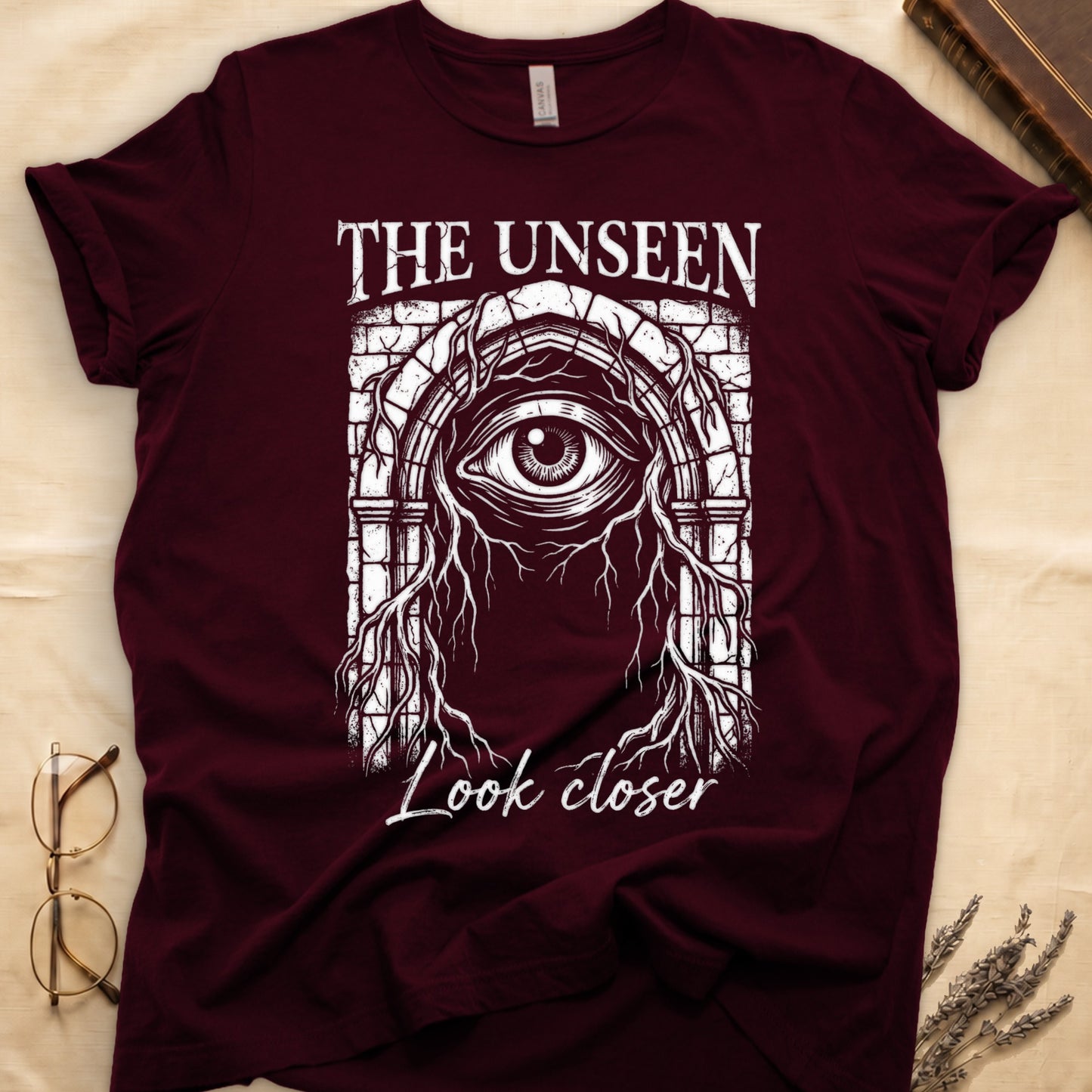 Maroon tee with Gothic stone arch, open eye, roots trailing, "THE UNSEEN" top, "Look closer" cursive below.