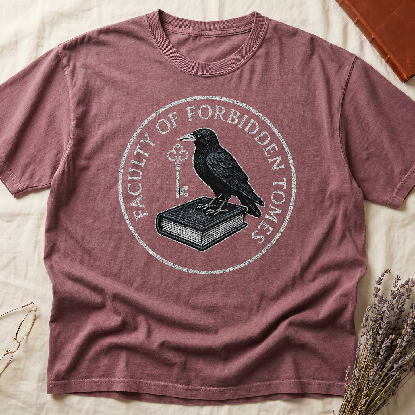 Blue Jean tee with circular badge, raven on closed book holding key, "FACULTY OF FORBIDDEN TOMES" curved around in cream.