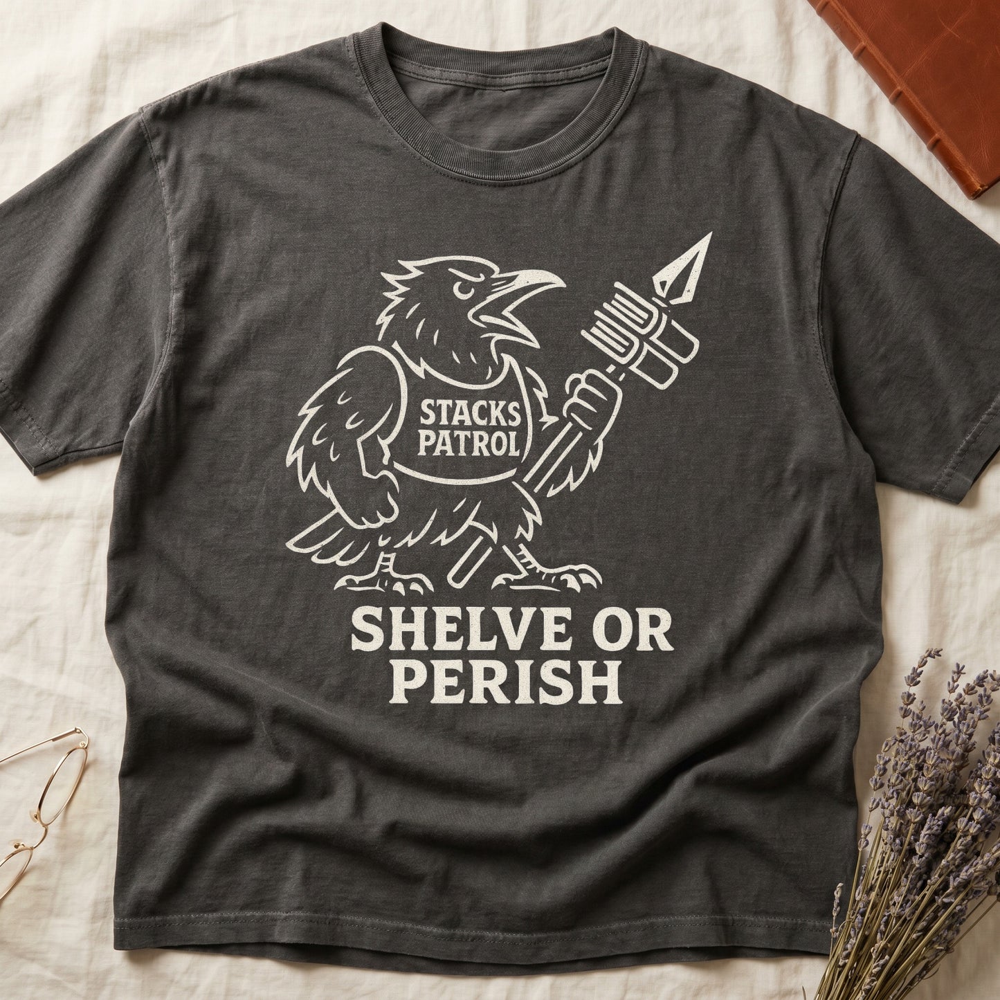Pepper tee with vintage raven mascot, "STACKS PATROL" sash, feather pen aloft, "SHELVE OR PERISH" bold serif below.