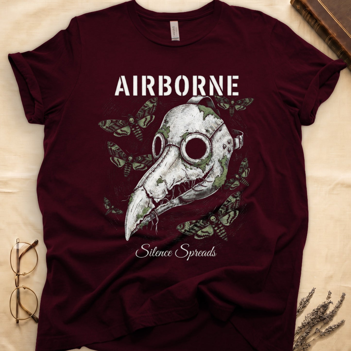 Maroon tee with plague doctor mask in cream linework, death's-head moths, "AIRBORNE" top, "Silence Spreads" cursive below.