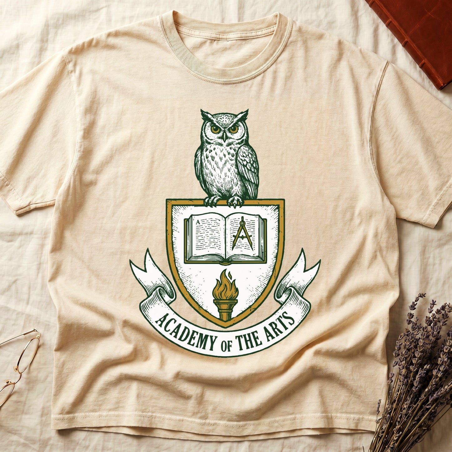 Ivory tee with heraldic shield crest, owl perched on top, open book and dividers center, "ACADEMY OF THE ARTS" on a banner.