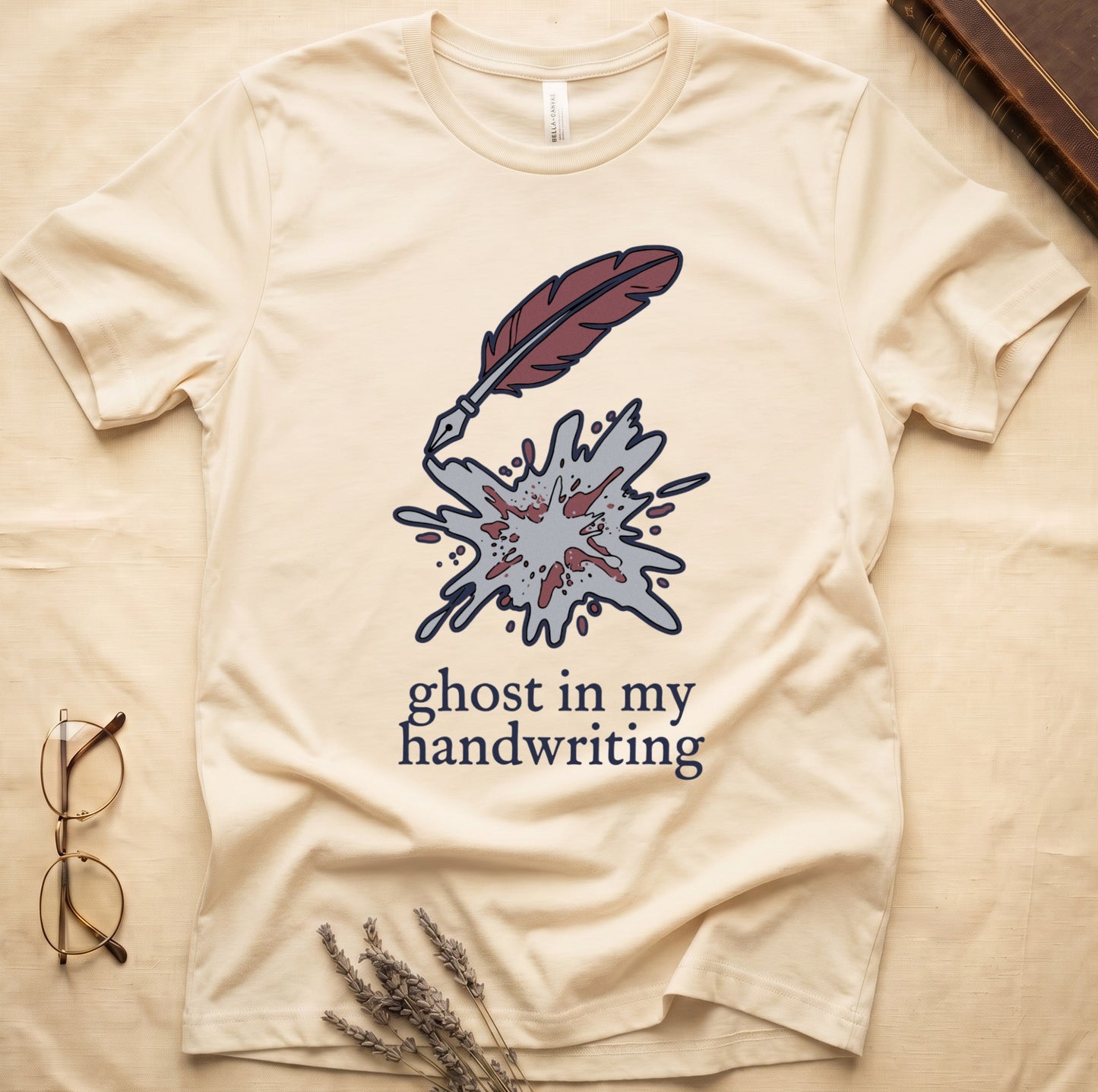 Natural tee with burgundy quill pen and grey ink splatter, "ghost in my handwriting" cream serif below.