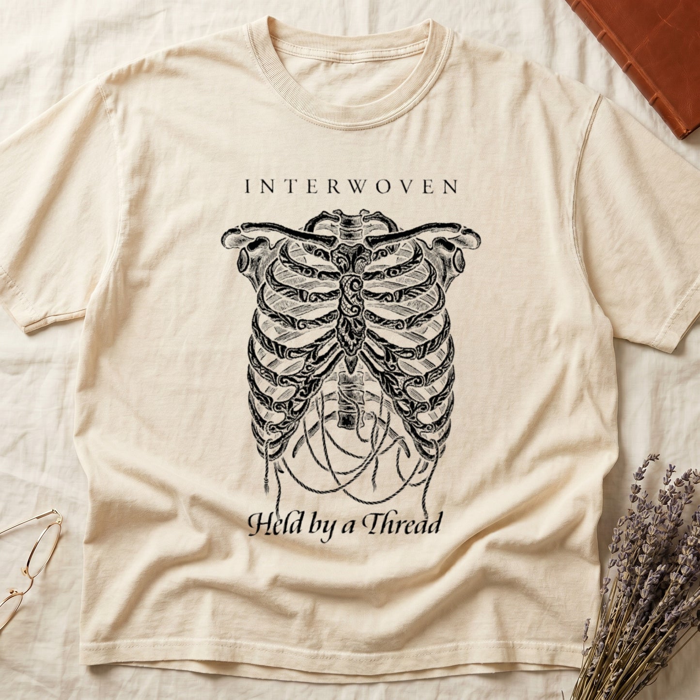 Ivory tee with cream skeletal ribcage suspended by trailing threads, "Held by a Thread" in serif on plate below.