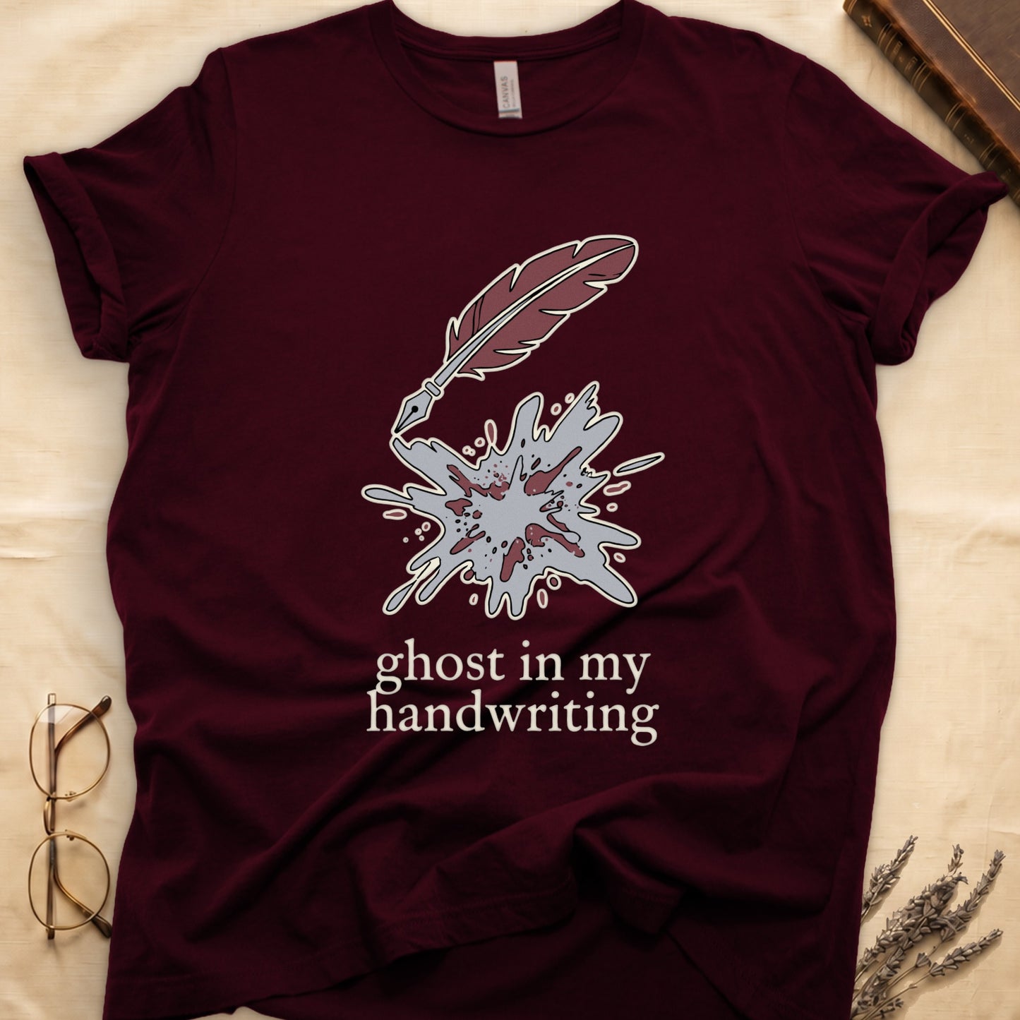 Maroon tee with burgundy quill pen and grey ink splatter, "ghost in my handwriting" cream serif below.