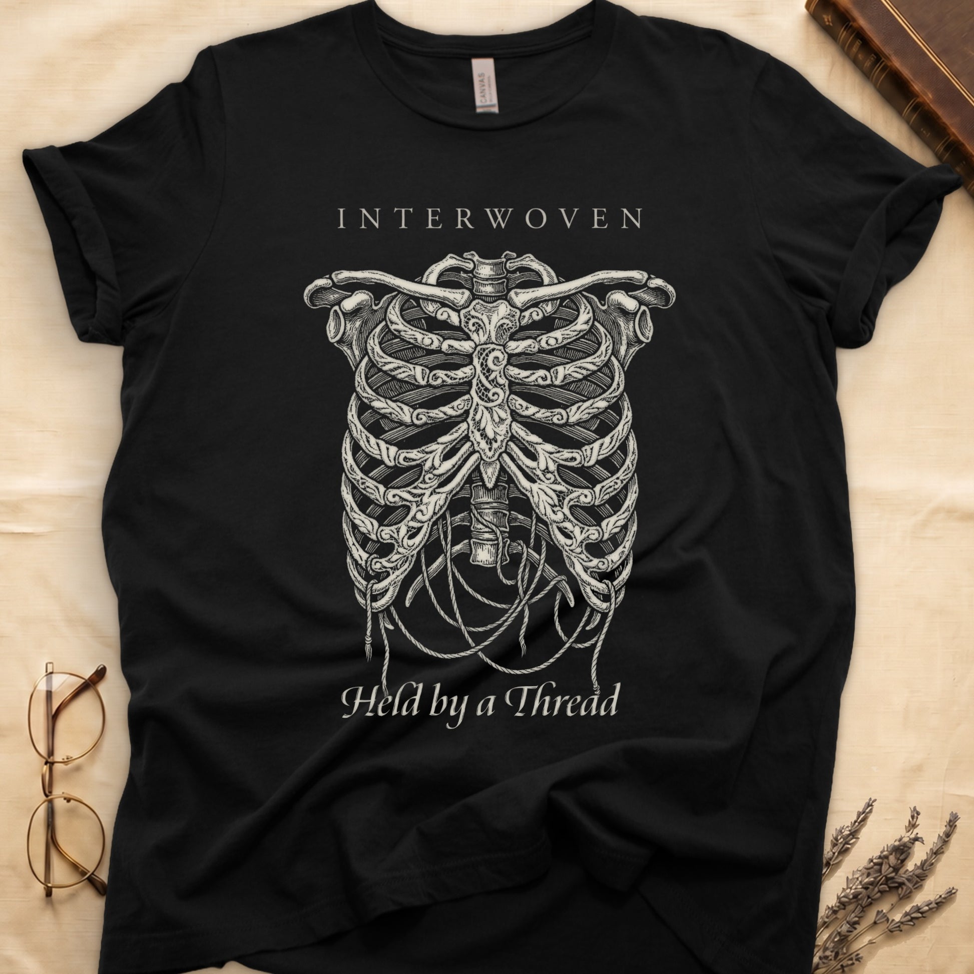 Black tee with cream skeletal ribcage suspended by trailing threads, "Held by a Thread" in serif on plate below.