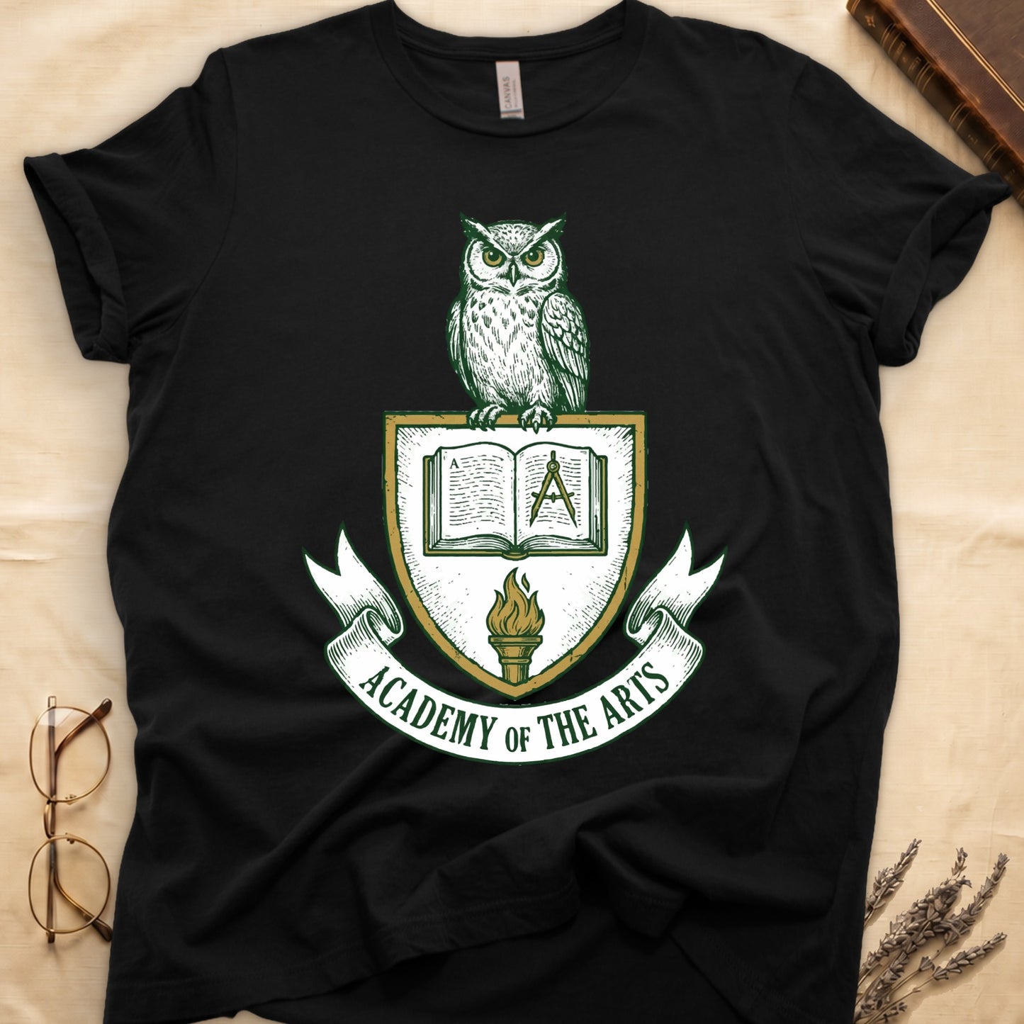 Black tee with heraldic shield crest, owl perched on top, open book and dividers center, "ACADEMY OF THE ARTS" on a banner.