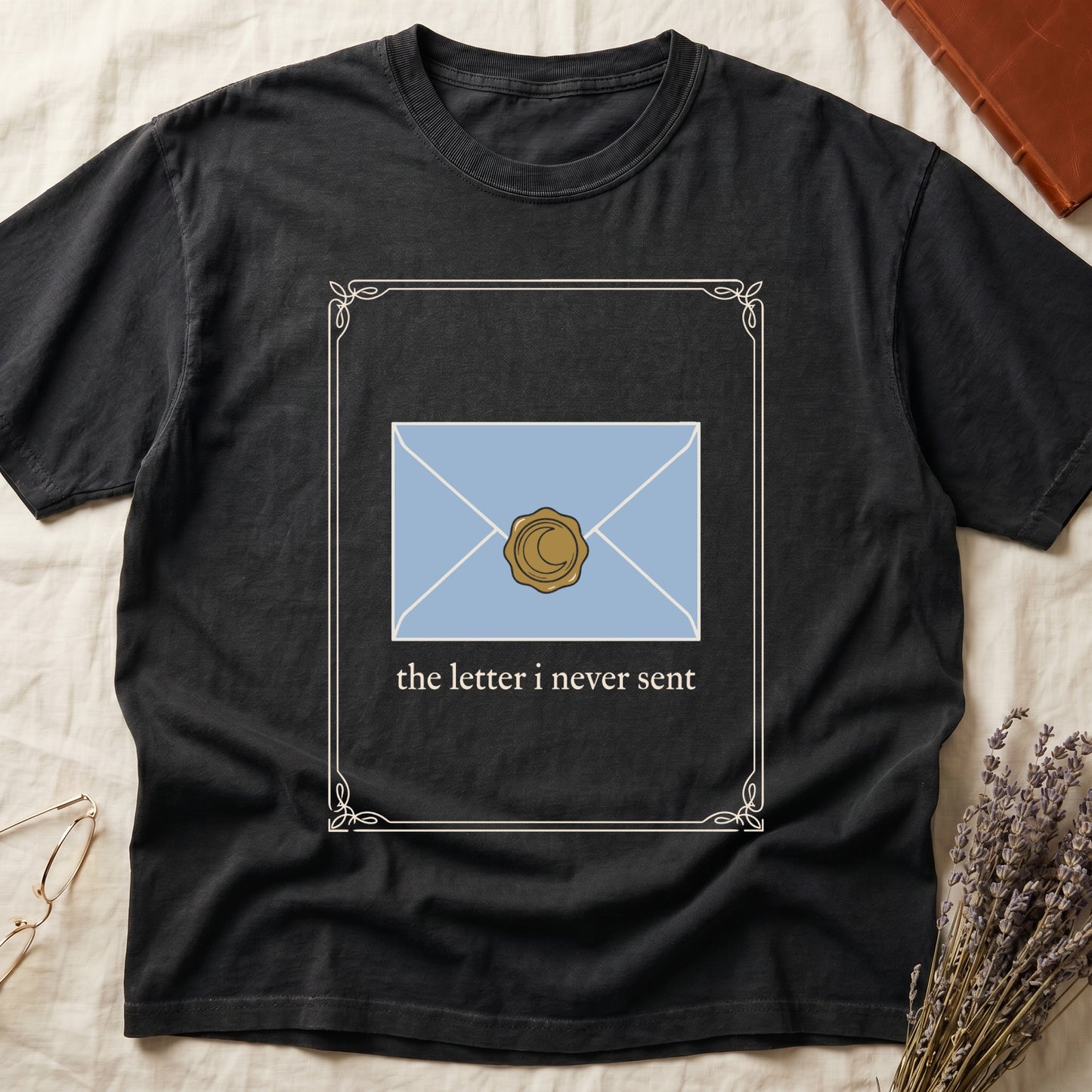 Black tee with pale blue envelope sealed with gold crescent moon wax seal, "the letter i never sent" cream serif below.