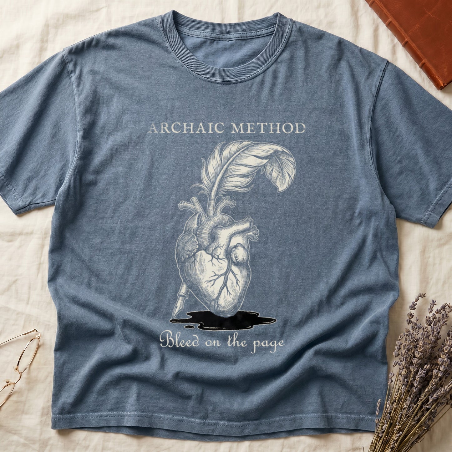 Blue Jean tee with anatomical heart and quill emerging in cream linework, "ARCHAIC METHOD" top, "Bleed on the page" below.