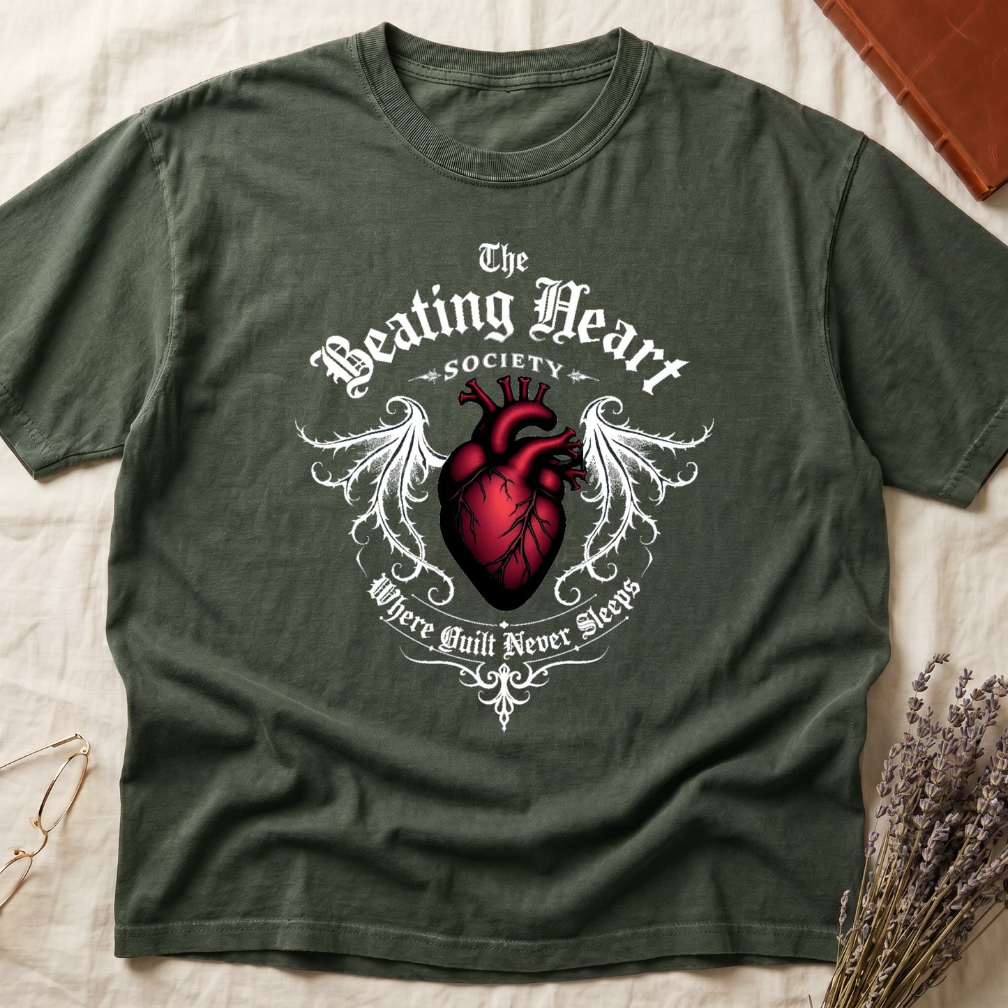 Green tee with anatomical red heart, gothic wings, "The Beating Heart SOCIETY," "Where Guilt Never Sleeps" below.