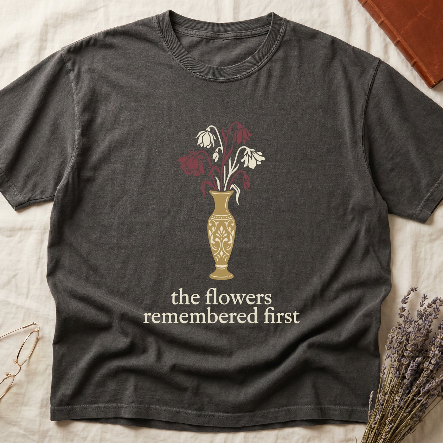 Pepper tee with gold ornate vase, wilting cream lilies and dark red roses, "the flowers remembered first" in cream serif.