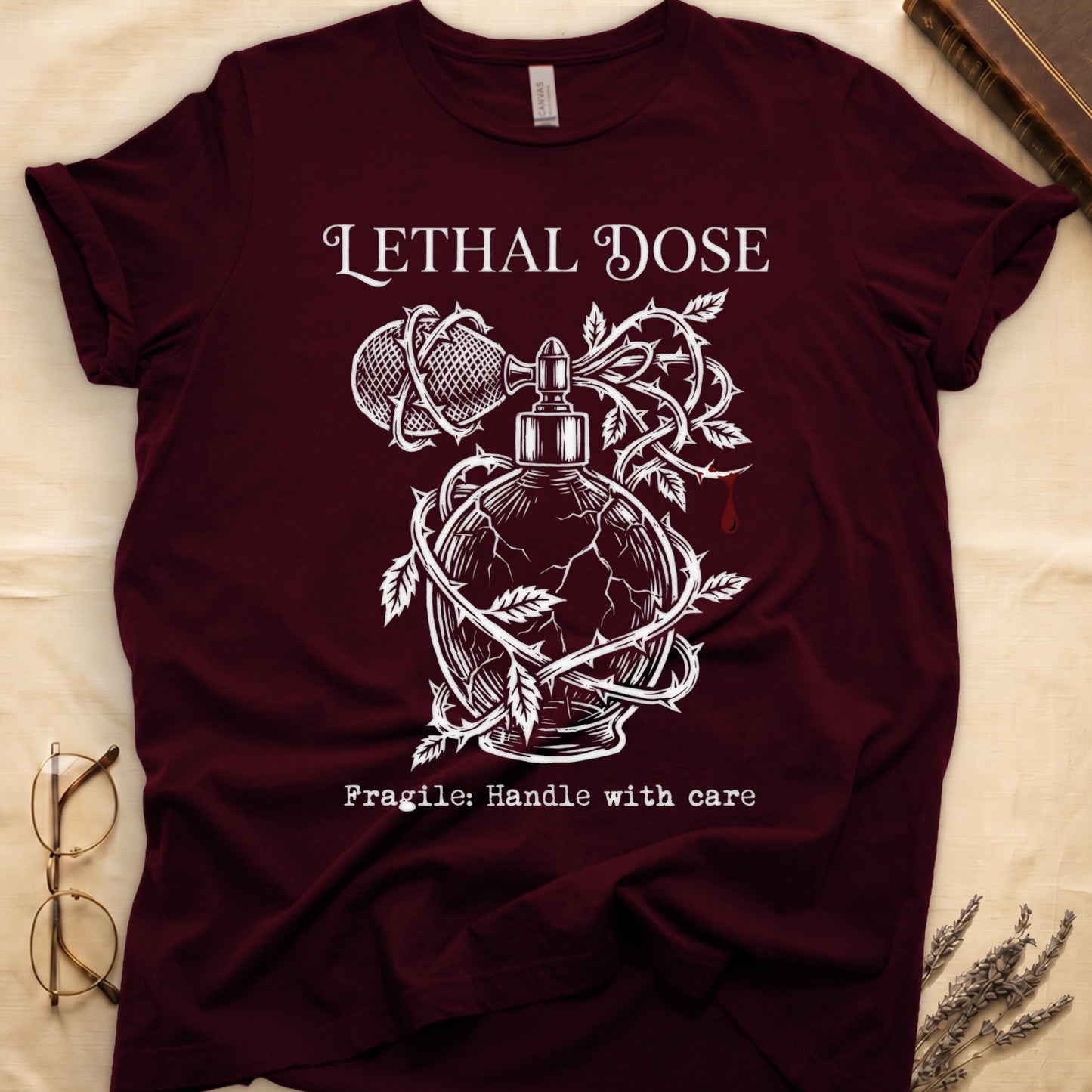 Maroon tee with cracked perfume bottle wrapped in thorns, blood drop, "LETHAL DOSE" top, "Fragile: Handle with care" below.