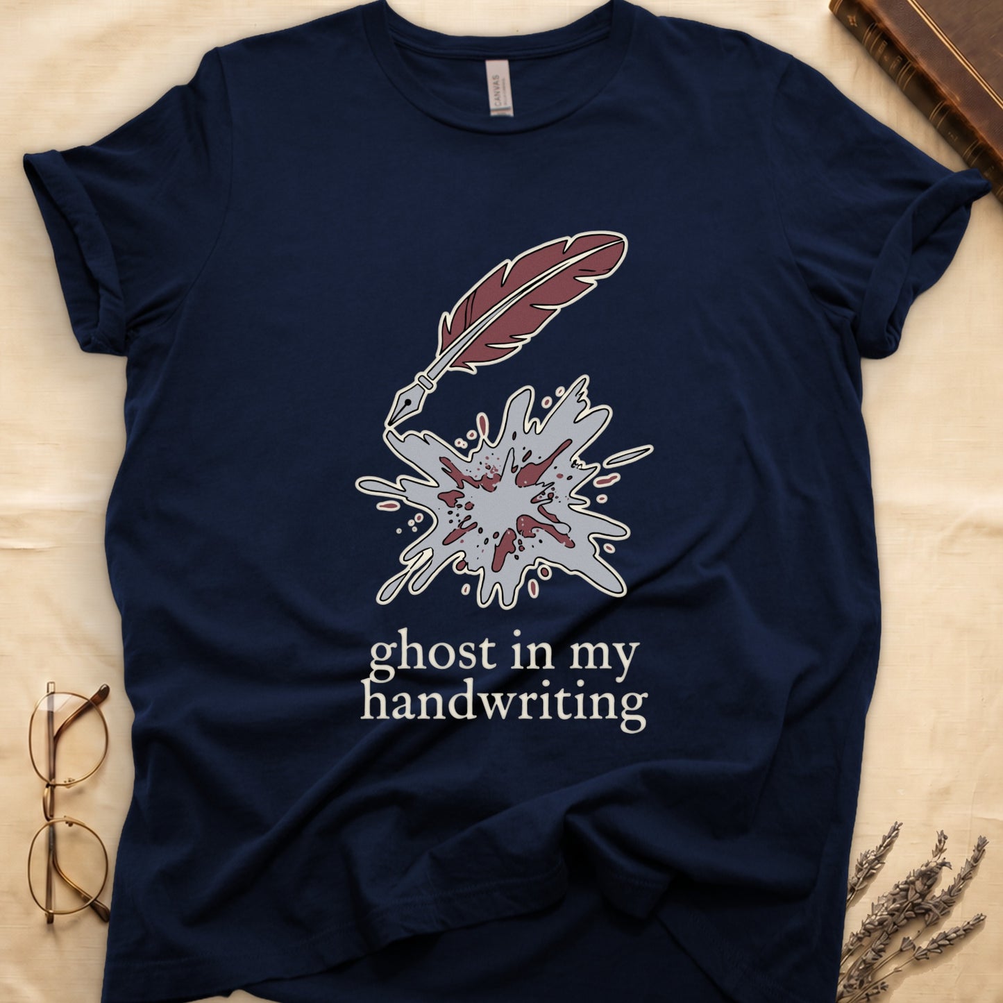 Navy tee with burgundy quill pen and grey ink splatter, "ghost in my handwriting" cream serif below.